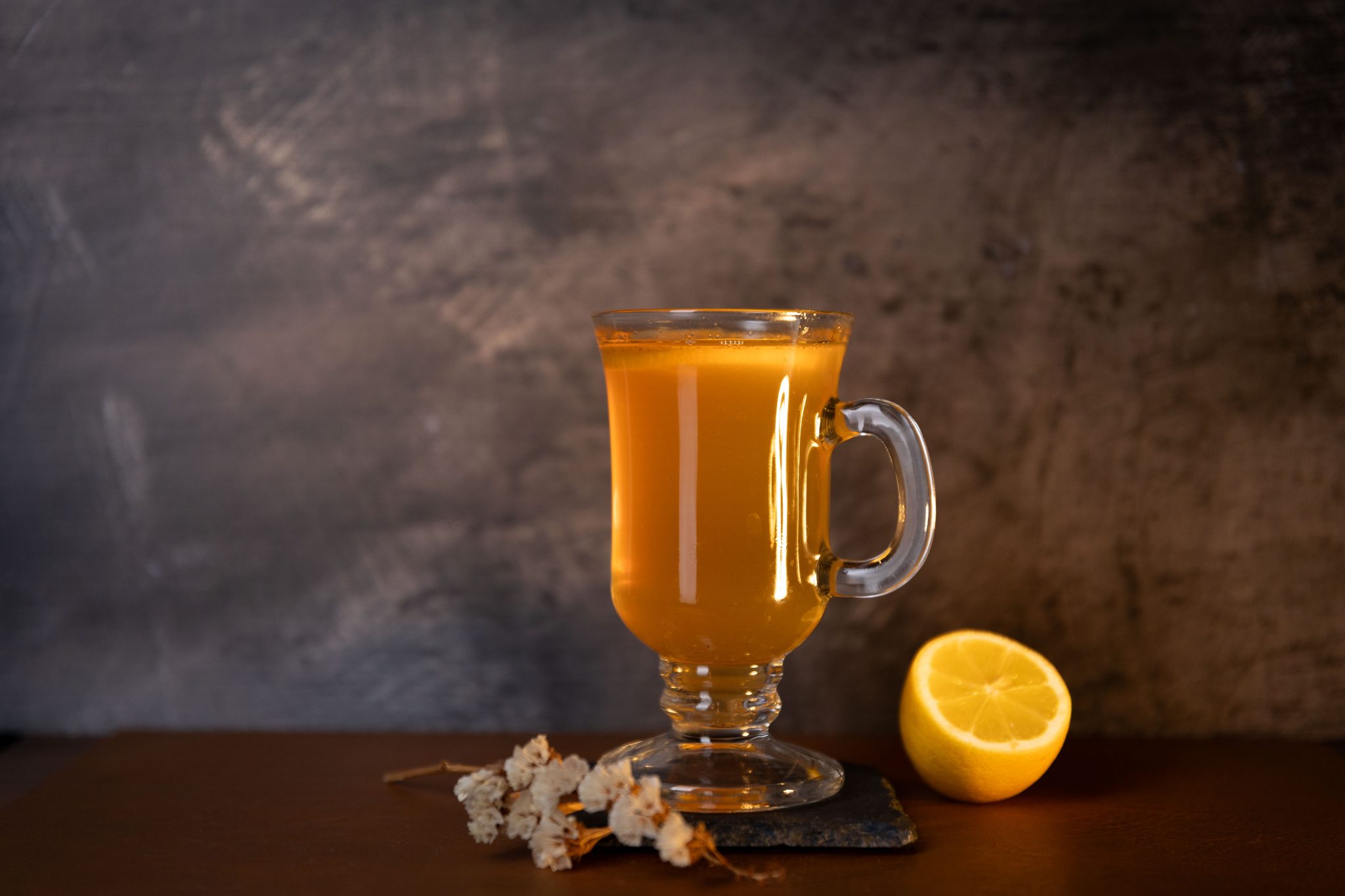 A side shot of a Fallen Leaf cocktail in a mug on a black stone coaster placed on a brown surface surrounded by half a lemon and flower sprig