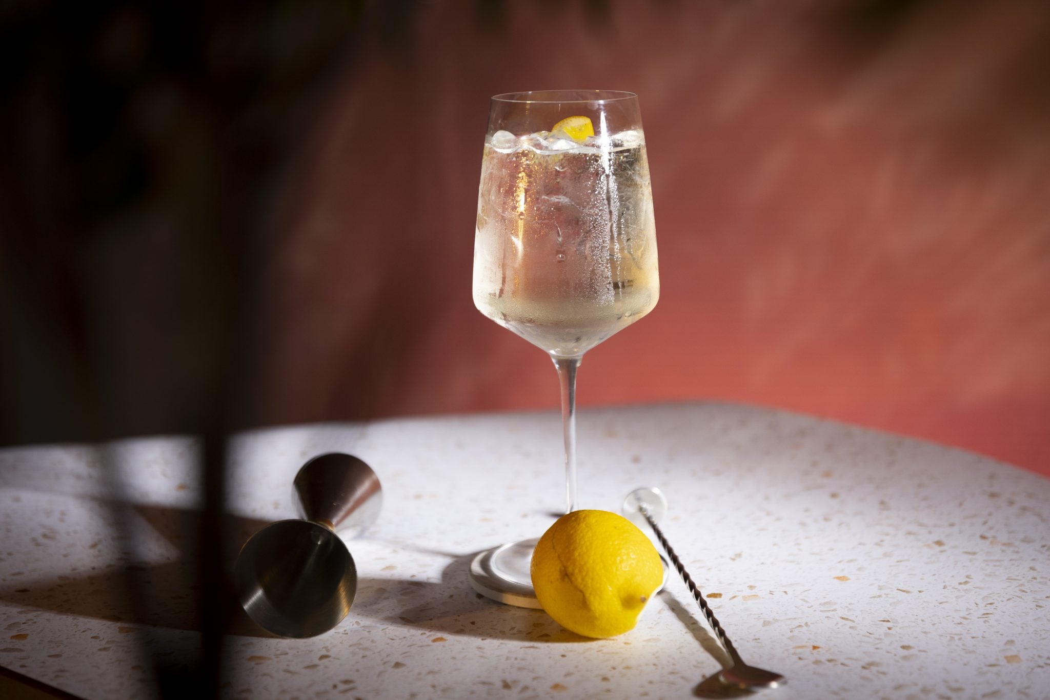 A side shot of a Gin Spritz cocktail in a wine glass on a white surface surrounded by a lemon, a jigger, and a bar spoon