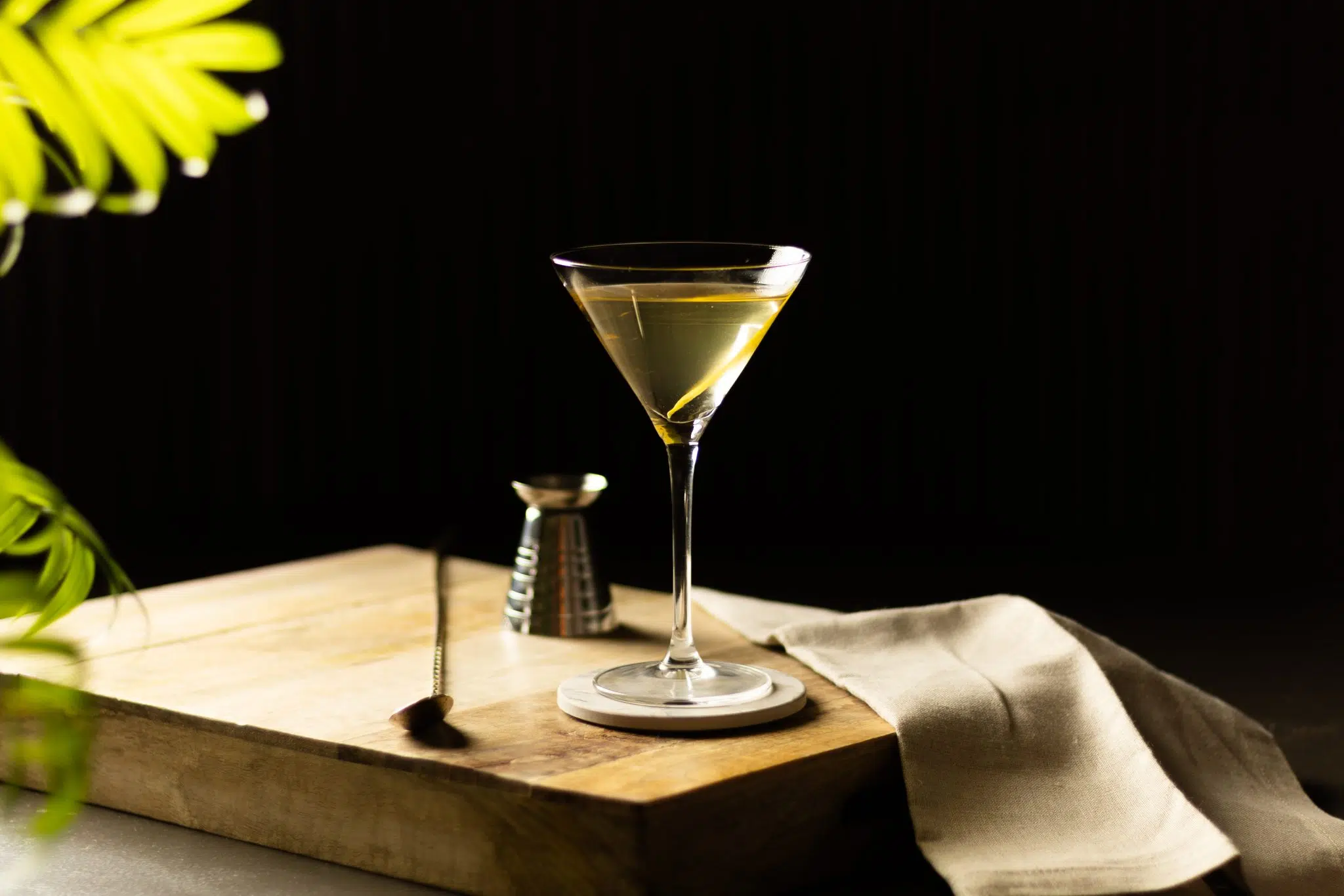 A side shot of a 50/50 Martini cocktail in a martini glass on a white coaster placed on a wooden board surrounded by a jigger, a bar spoon, and a white cloth, in front of a black background