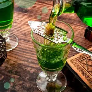 Absinthe poured over a brown sugar cube on a knife with holes over a glass placed on a dark brown wooden table.