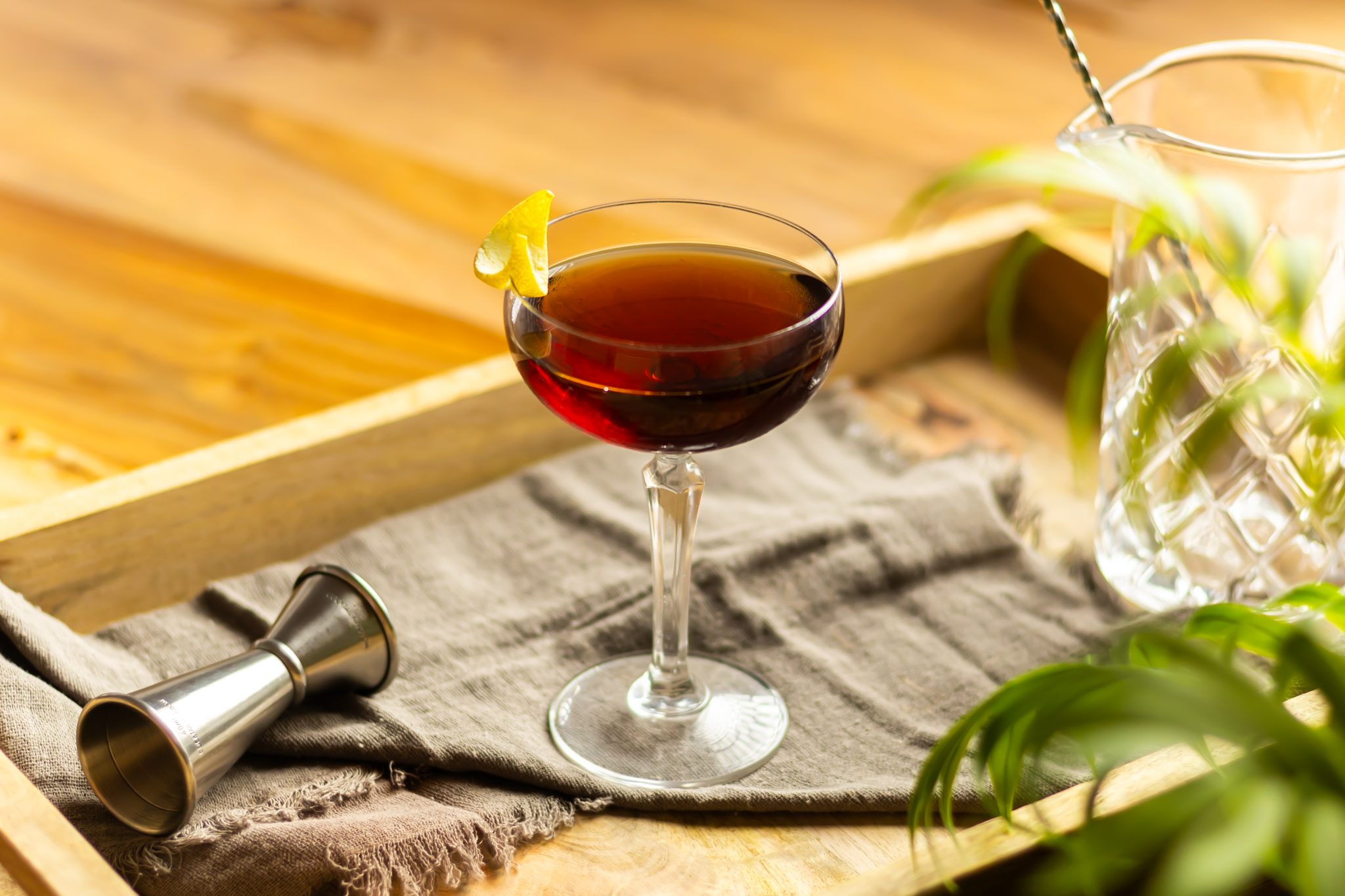 A side shot of an Affinity cocktail in a cocktail glass on a wooden tray on a wooden table surrounded by a grey cloth, and a jigger