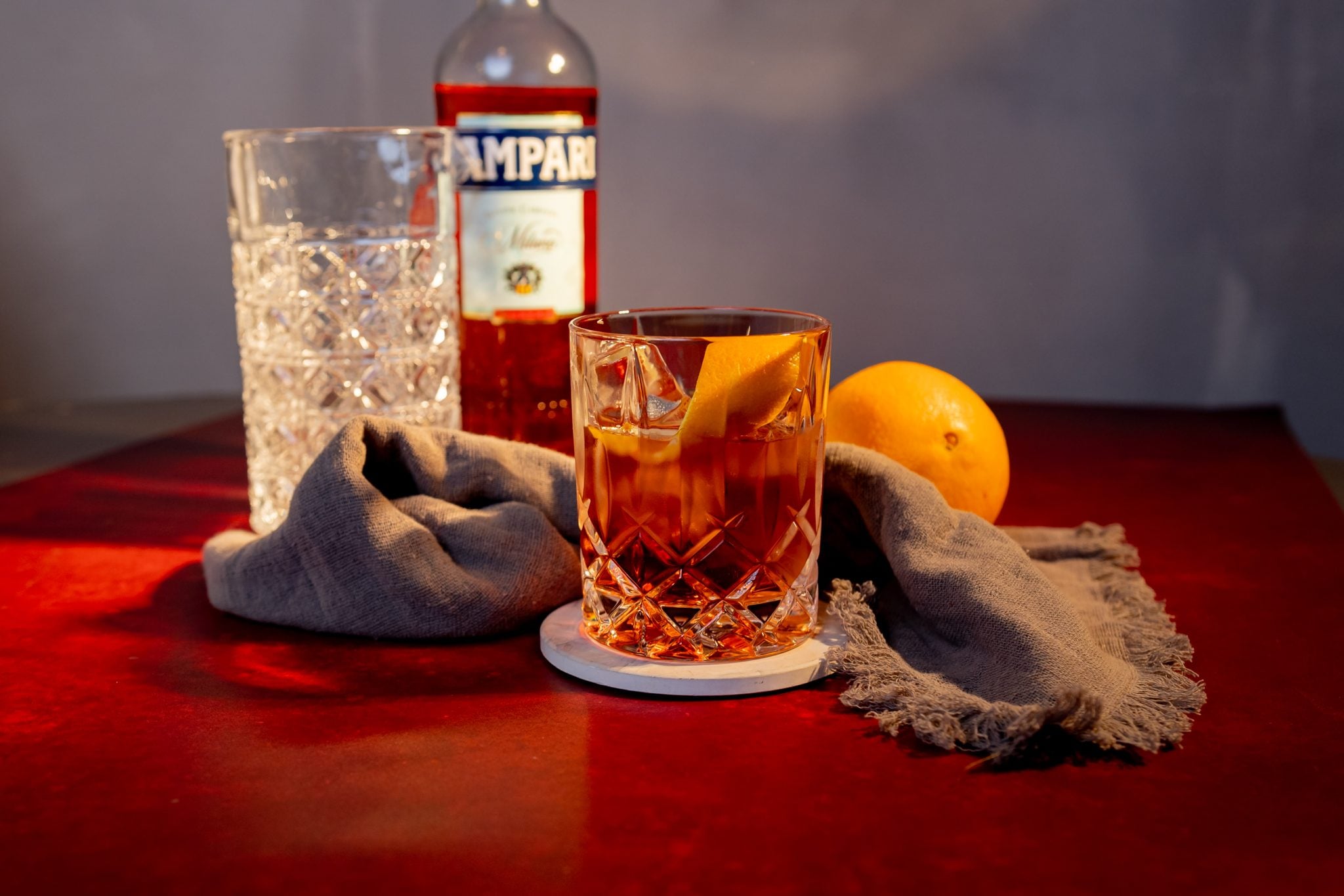 A side shot of a Agavoni cocktail in an old fashioned glass on a white coaster placed on a wooden surface surrounded by a grey cloth, a mixing glass, a Campari botle and an orange