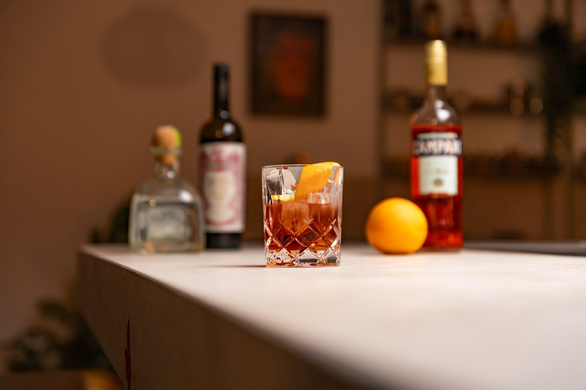 Tequila, Campari, sweet vermouth and orange laid out on a white bar table