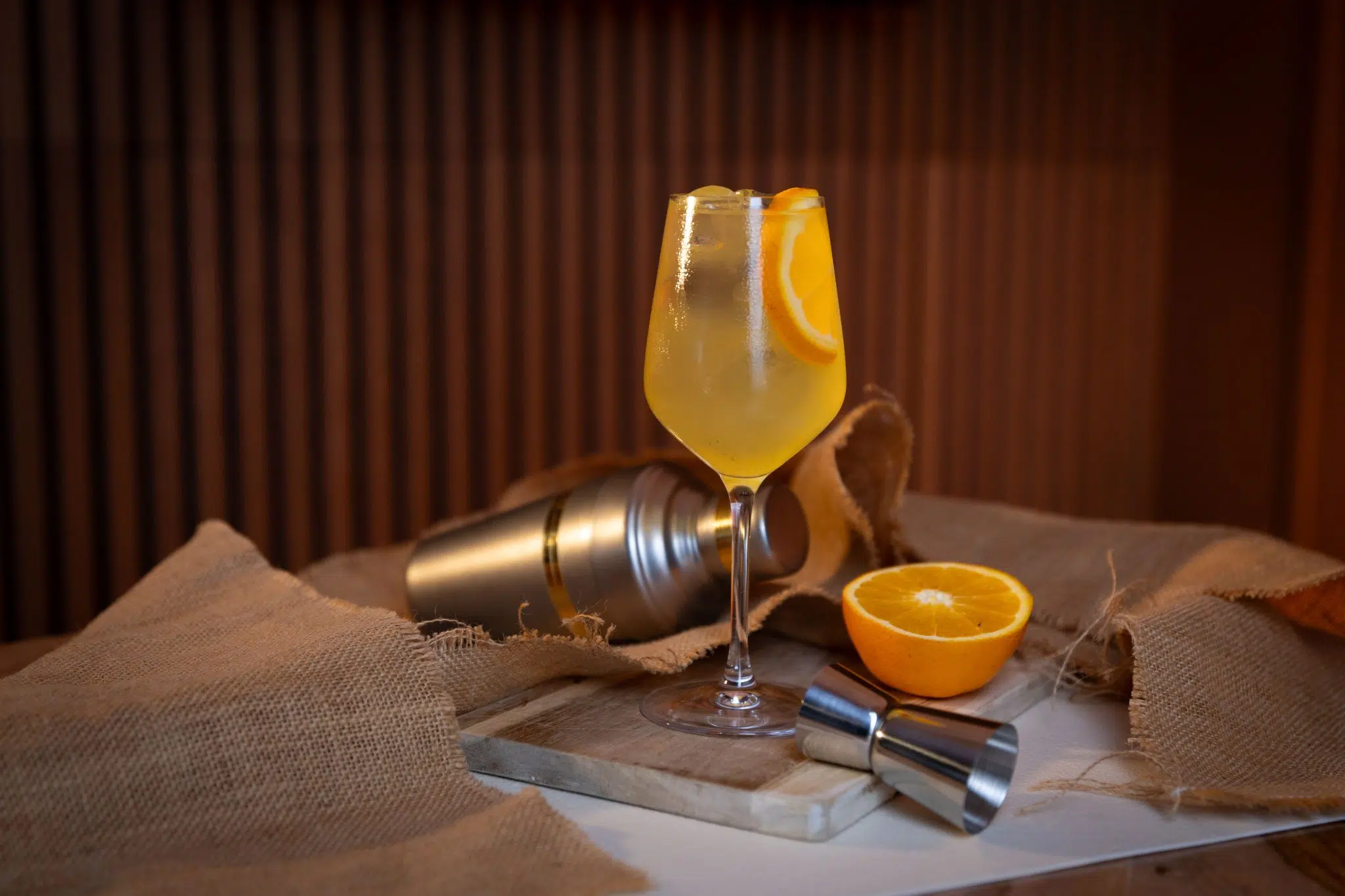 A side shot of an Agua de Valencia cocktail in a wine glass on a wooden board placed on a white surfacee surrounded by a jigger, a cocktail shaker, an orange and a brown cloth