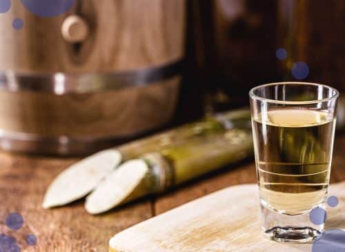 A glass with aguardiente on a light brown wooden tray placed on a dark brown wooden surface, with two sugar cane and a barrel behind.