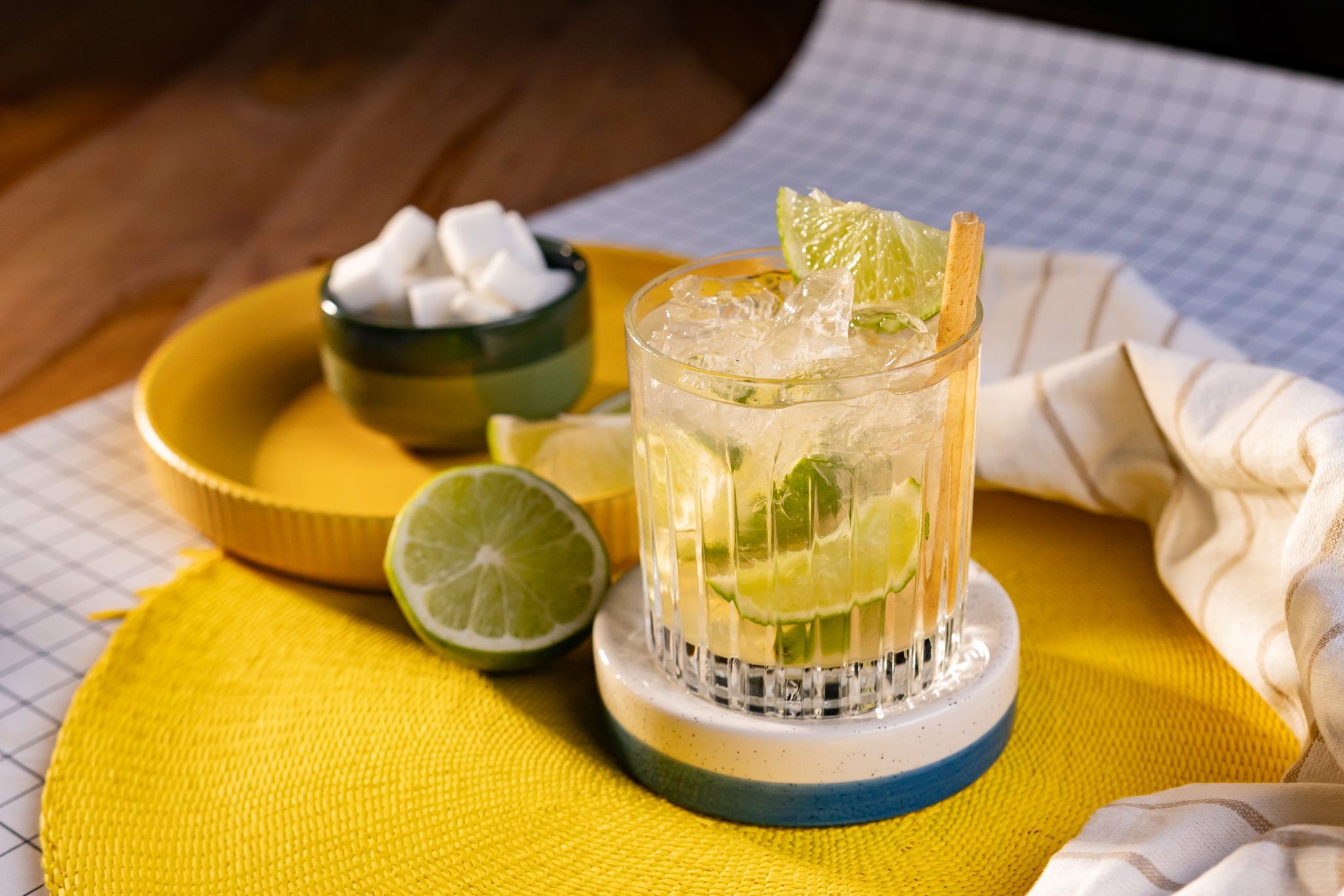 A side shot of an Aguardiente Caipirinha cocktail in an old fashioned glass on a white and blue coaster placed on a yellow tray surrounded by half a lime, and a green bowl with sugar cubes