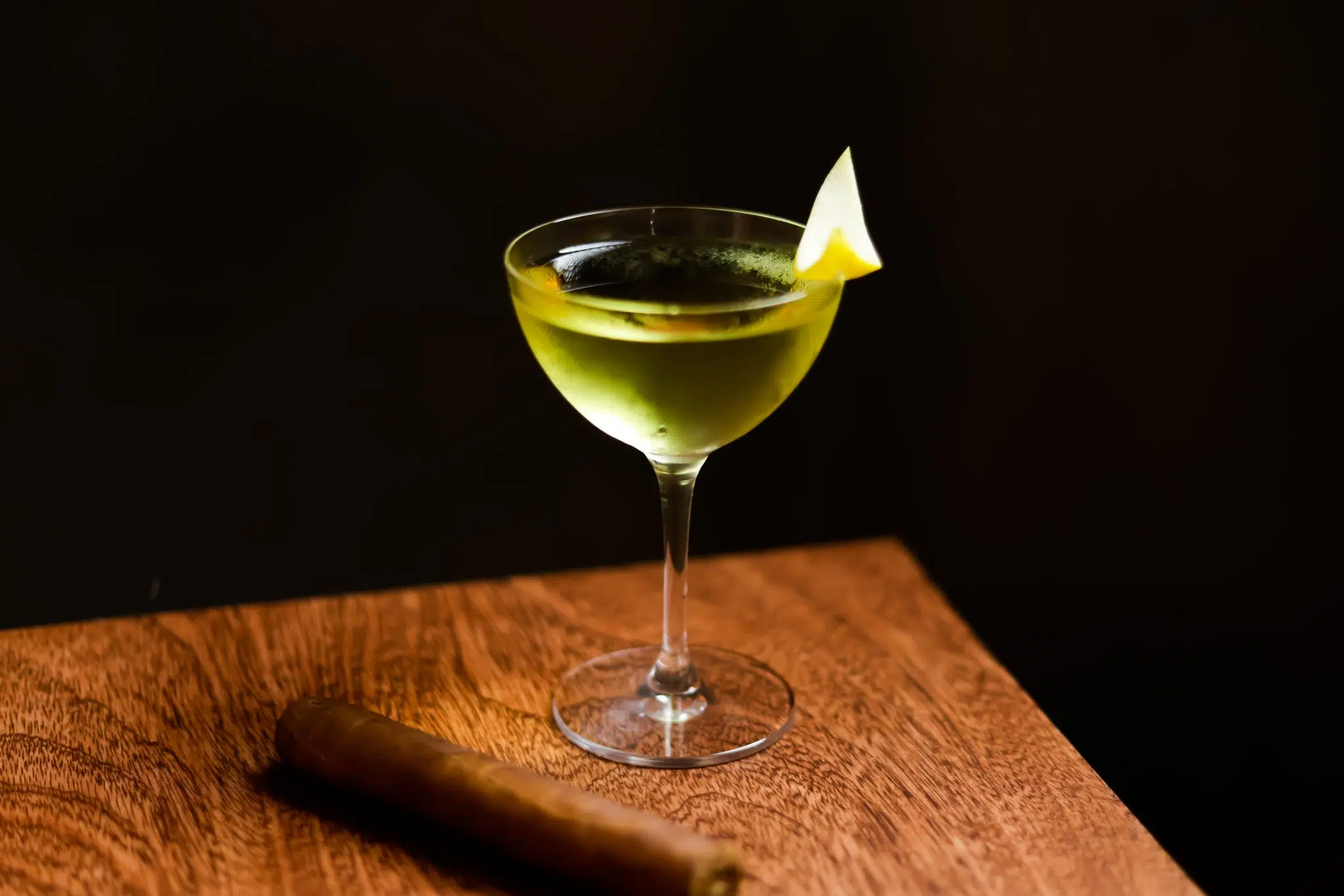 A side shot of an Alaska cocktail in a coupe glass on a wooden table with a cigar on the side and a black background