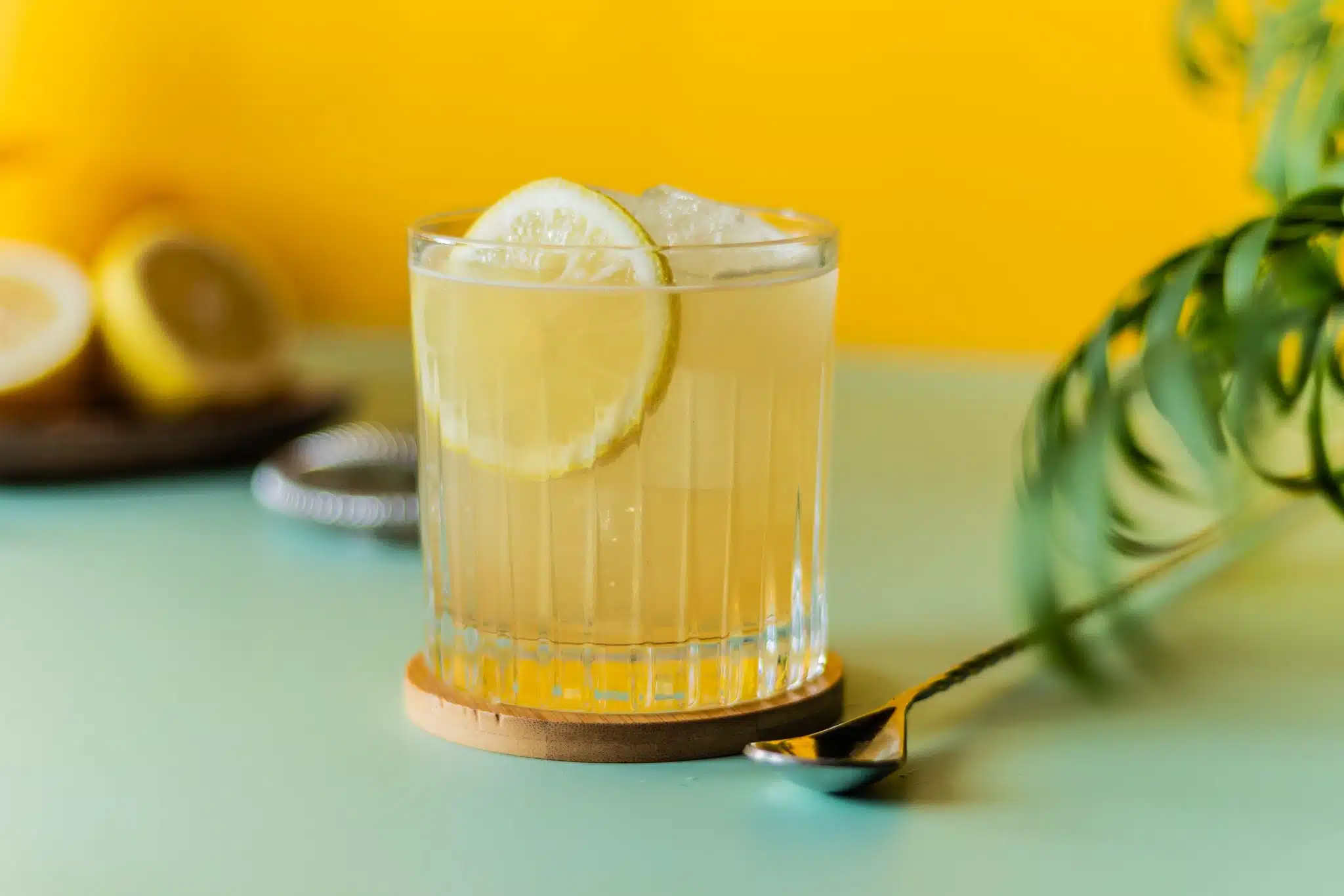 A side shot of an Amaretto Sour cocktail in an old fashioned glass on a wooden coaster with a bar spoon on the side placed on a turquoise table and in front of a yellow wall..