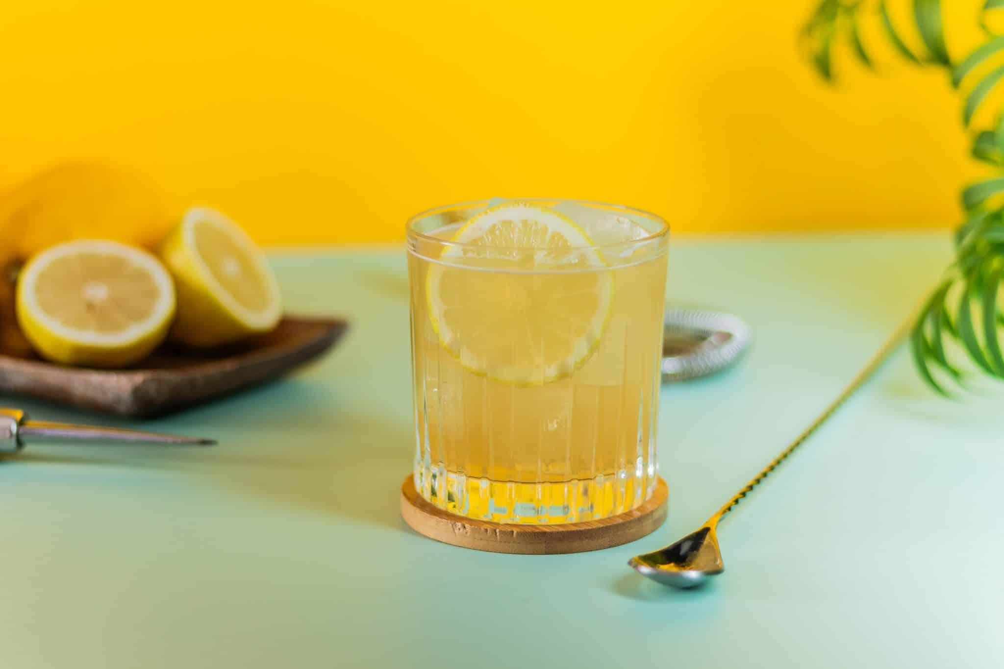 A side shot of an Amaretto Sour cocktail in an old fashioned glass on a wooden coaster with a bar spoon on the side placed on a turquoise table with two half lemons behind and in front of a yellow wall.