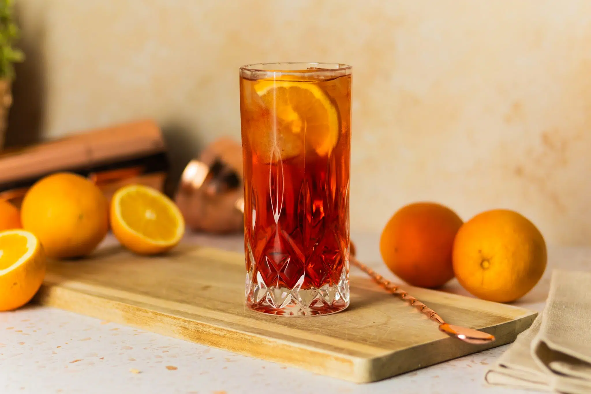 A side shot of an Americano cocktail in a highball glass on a wooden board placed on a white table surrounded by five oranges, a shaker, and a bar spoon.