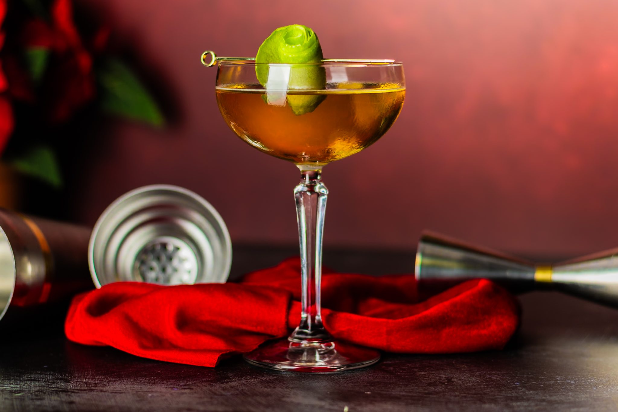 A side shot of an Angel Face cocktail in a cocktail glass on a black table surrounded by a shaker, a red cloth and a jigger.