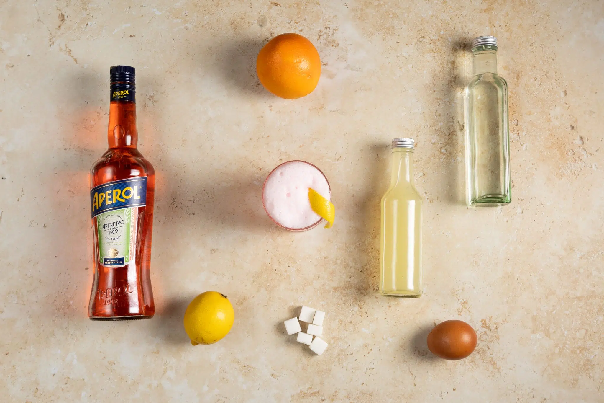 An Aperol Sour cocktail, shot from above, in an old fashioned glass on a beige surface surrounded by Aperol, lemon juice, simple syrup, lemon, orange, sugar cubes, and egg