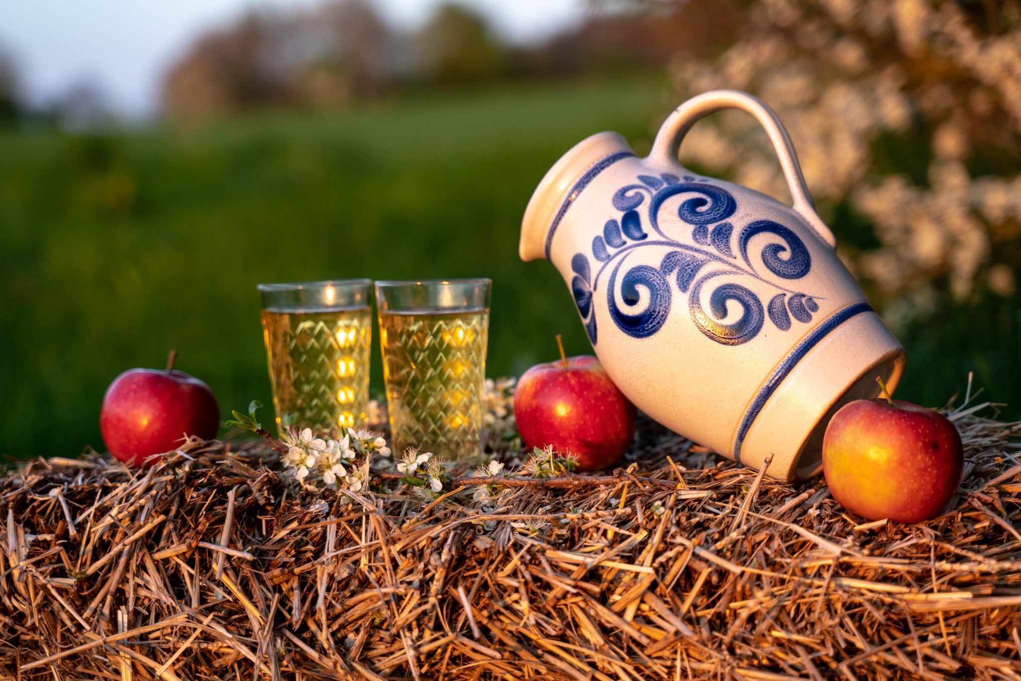 A shot two apfelwein drinks on a straw surface with three apples and a white and blue jug