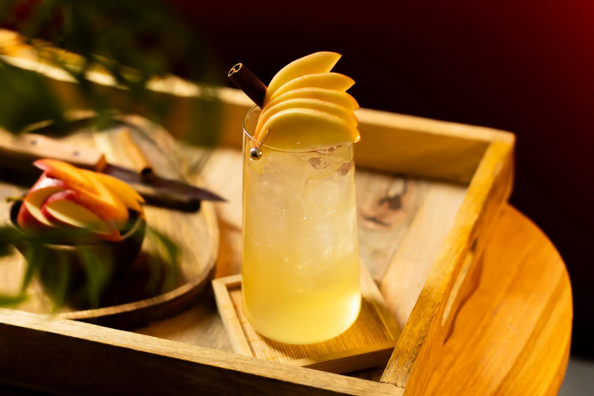 A side shot of a Apple Cider Mule cocktail in a highball glass on a wooden coaster placed on a wooden tray surrounded by apple slices, a wooden board and a knife.