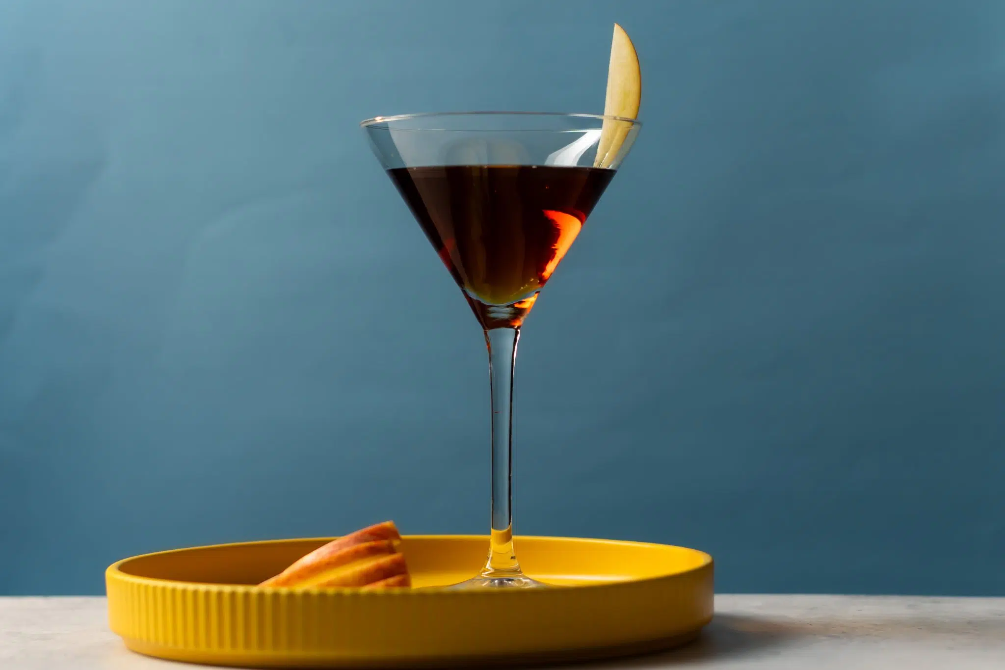A side shot of an Apple Manhattan cocktail in a martini glass on a yellow tray placed on a white surface with apple slice on a side, in front of a blue background