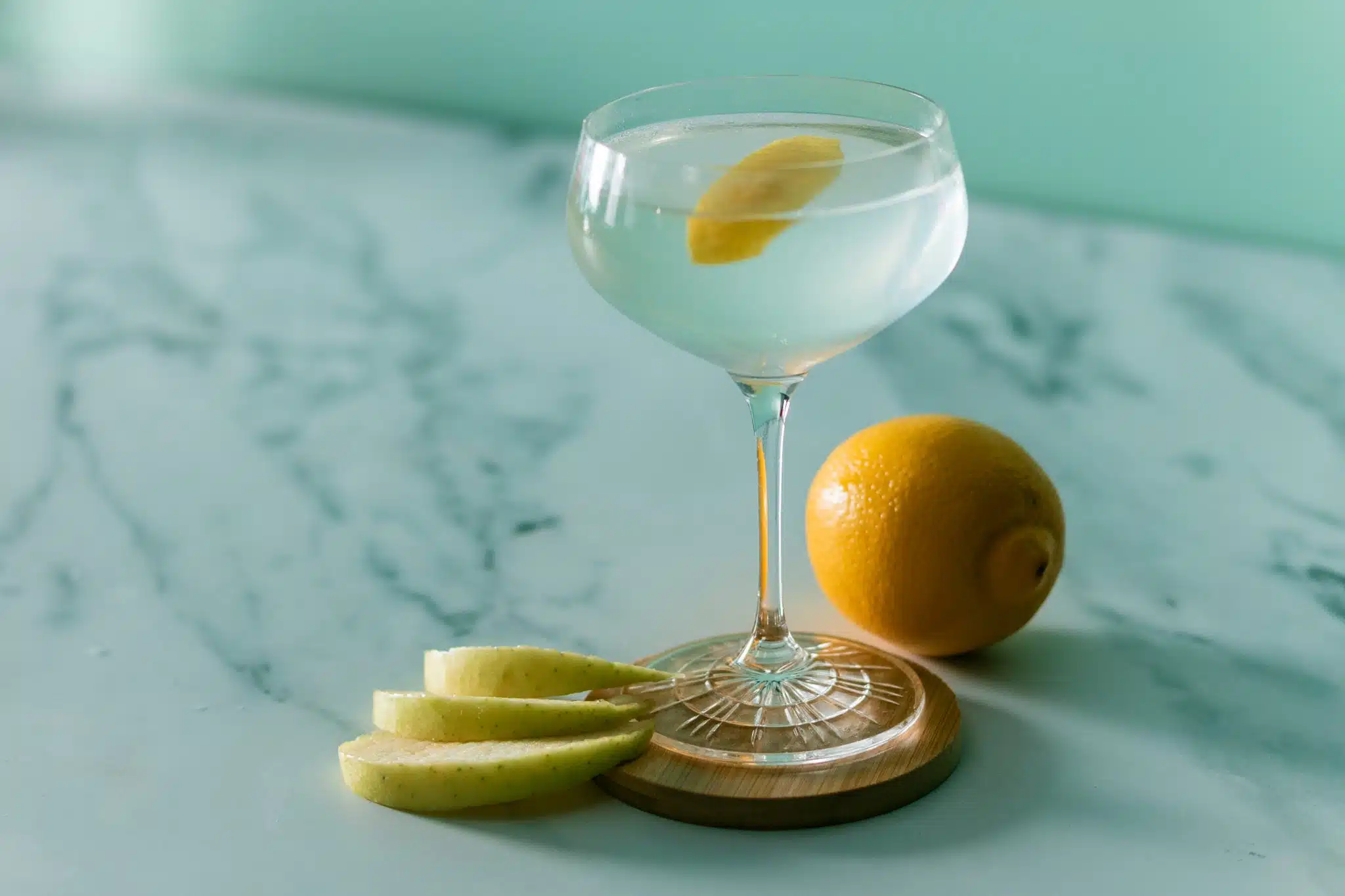 A side shot of an Apple Martini cocktail in a martini glass on a brown coaster with three apple slices and a lemon on the side, placed on a white marmol table.