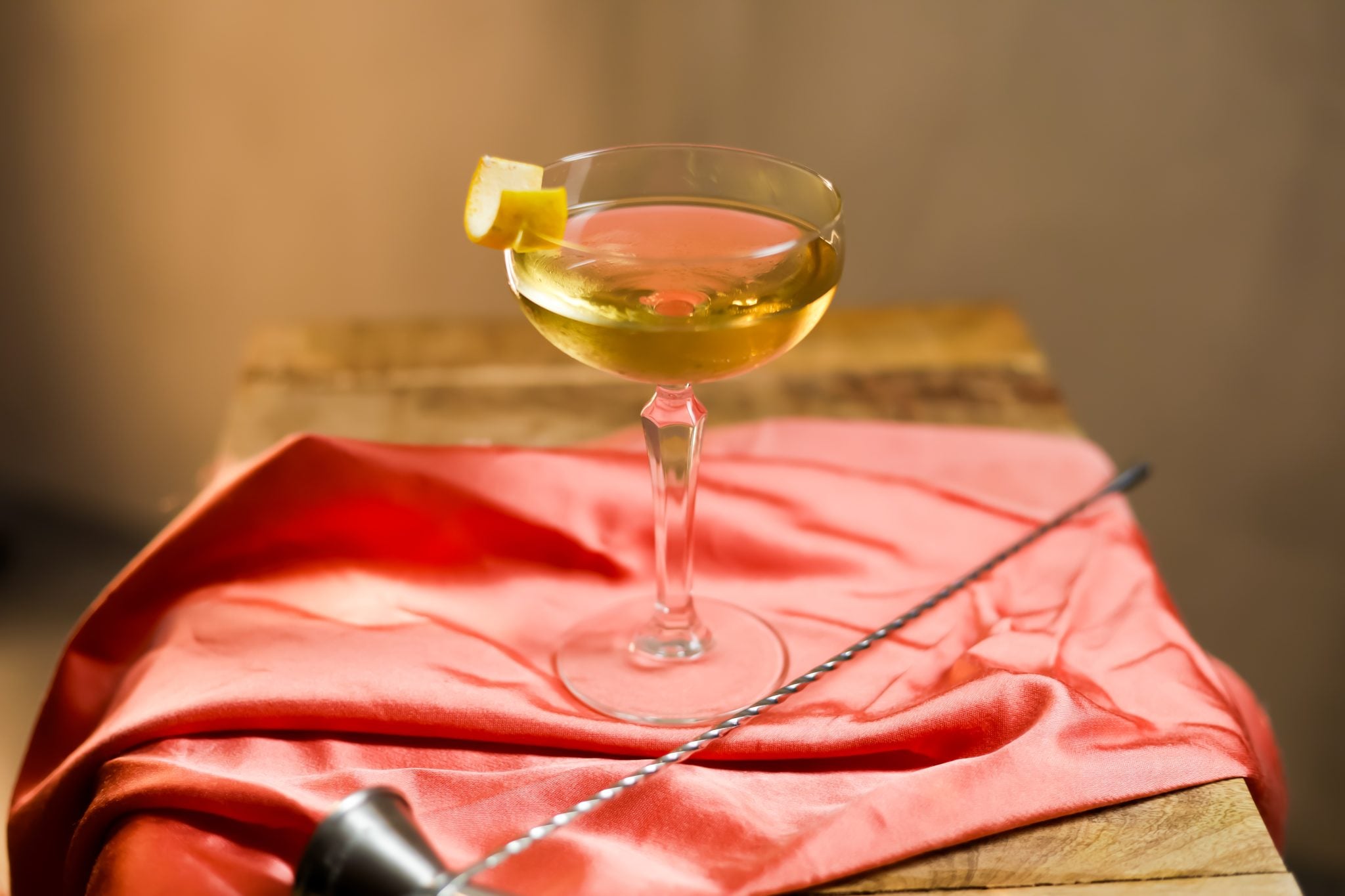 A side shot of an Astoria cocktail in a cocktail glass on a red cloth with a jigger and a bar spoon around