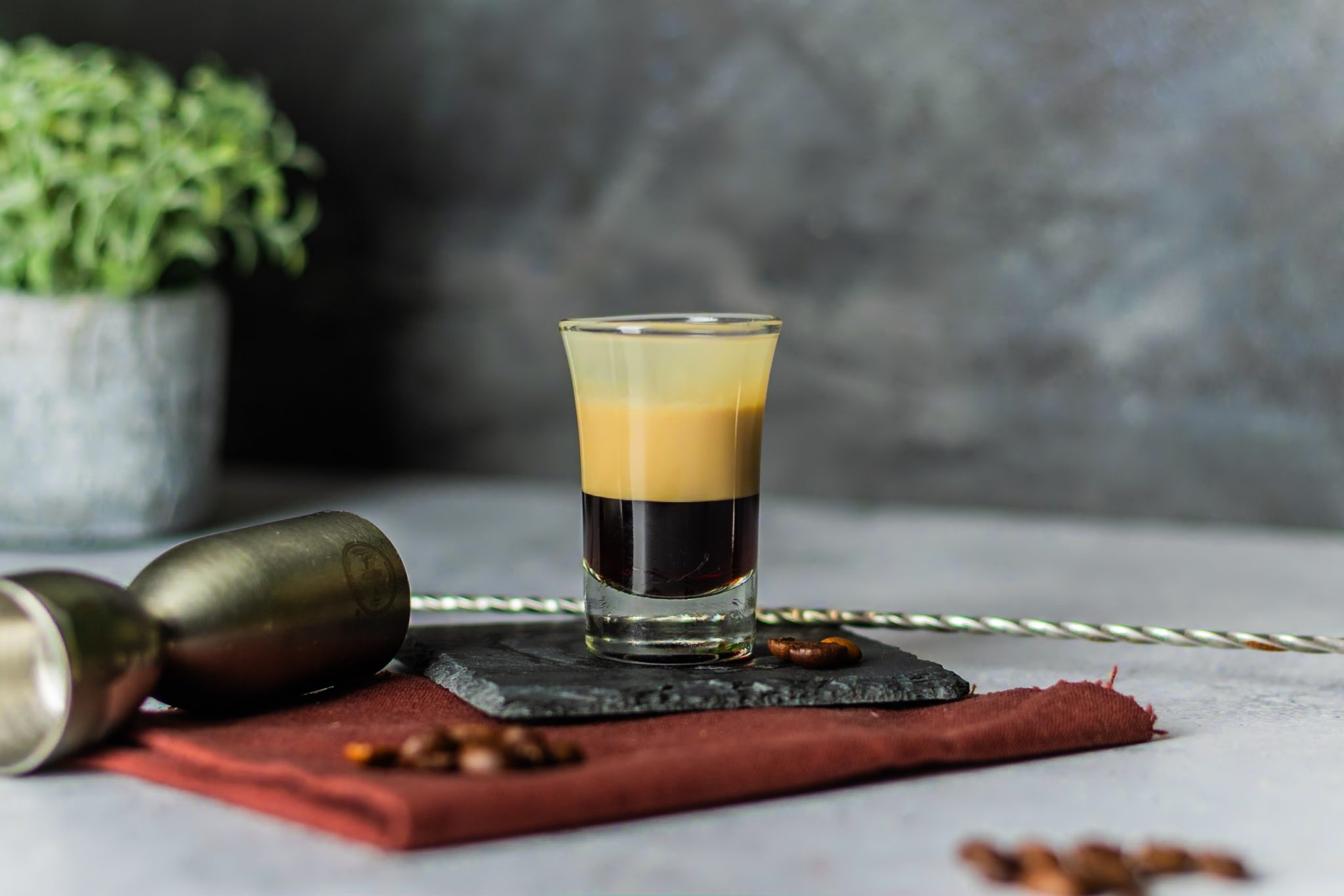 A side shot of a B-52 cocktail in a shot glass on a black stone coaster placed on a red cloth on a white table with a bar spoon, a jigger and coffee beans around, in front of a grey wall.