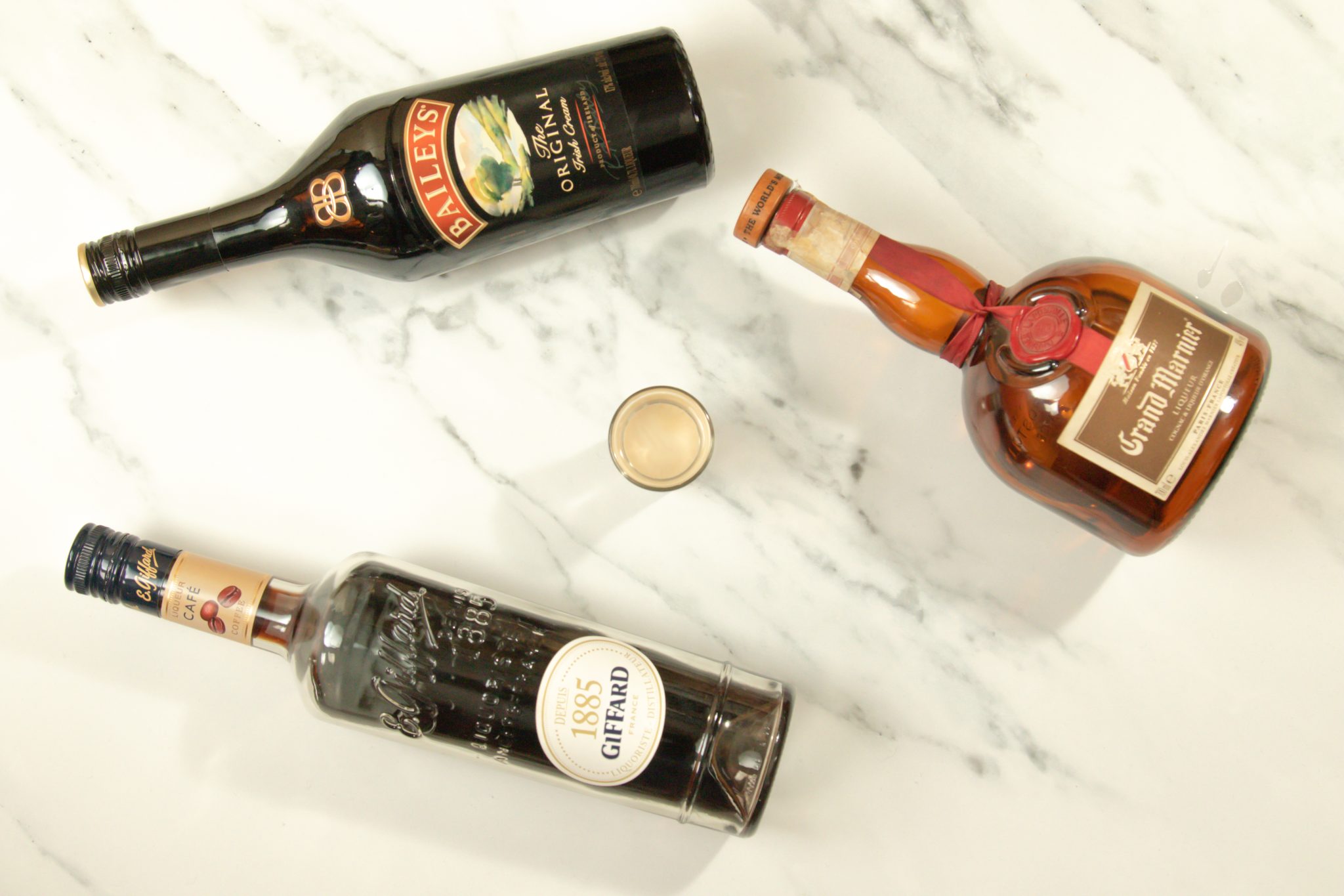 A B-52 cocktail, shot form above, on a white bar table surrounded by a Kahlúa bottle, a Bailey's Irish Cream bottle and a Grand Marnier bottle.
