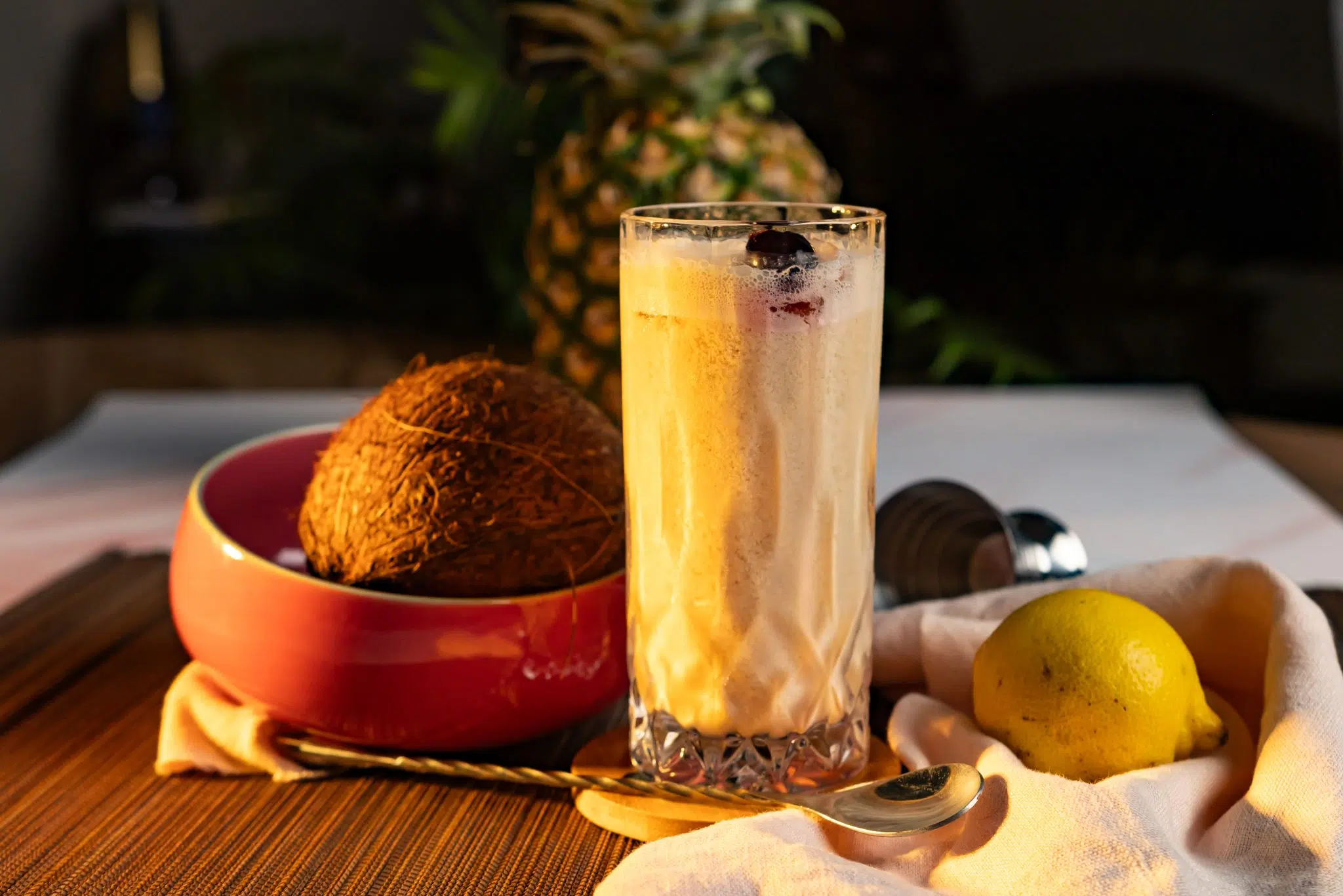 A side shot of a Bahama Papa cocktail in a highball glass on a wooden coaster placed on a brown placemat surrounded by a lemon, a white cloth, a jigger, a bar spoon, a pineapple and a coconut in a red bowl.