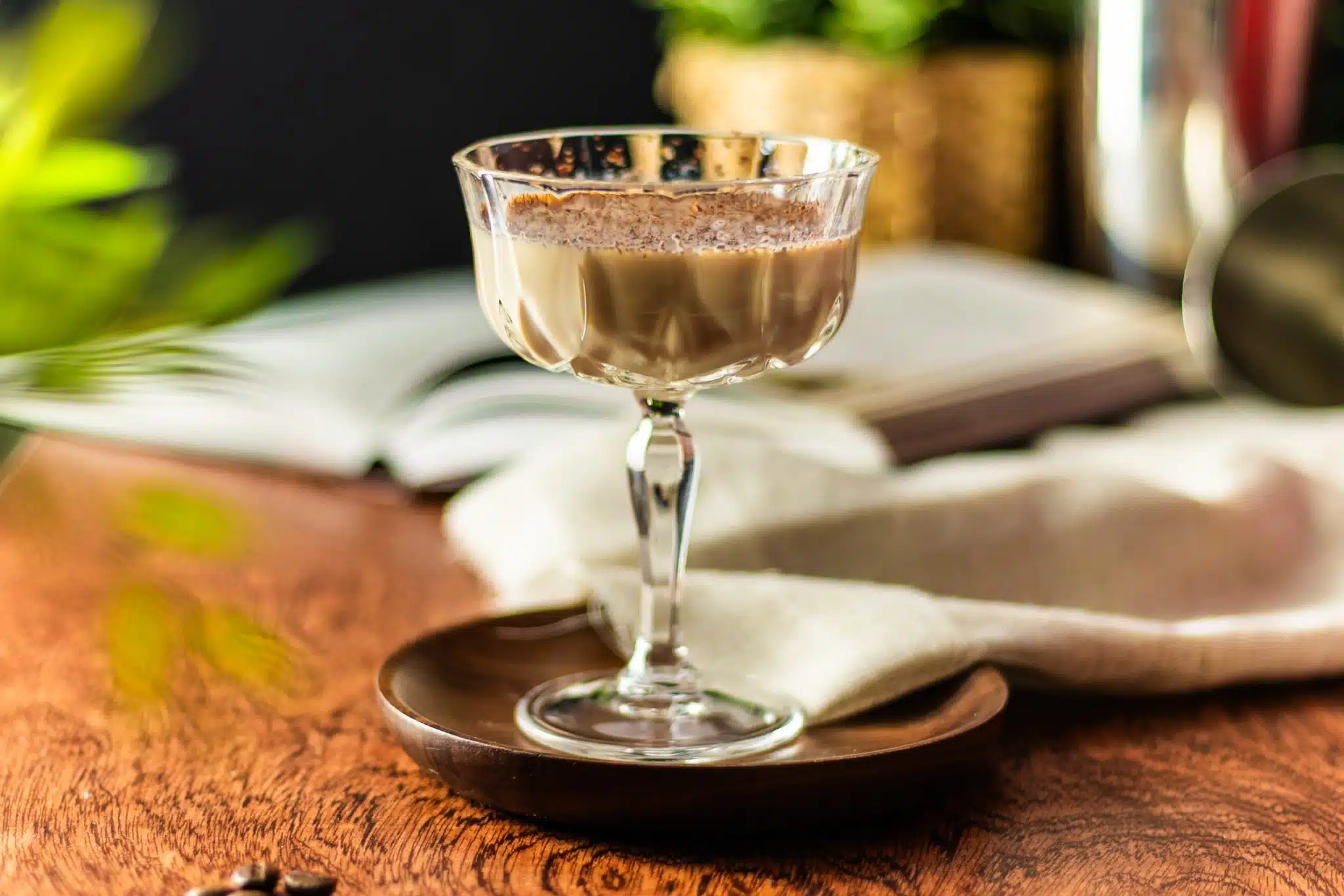 A side shot of a Bailey's Martini cocktail in a martini glass on a dark brown tray placed on a wooden table with a white cloth, a book and plat leaves around.