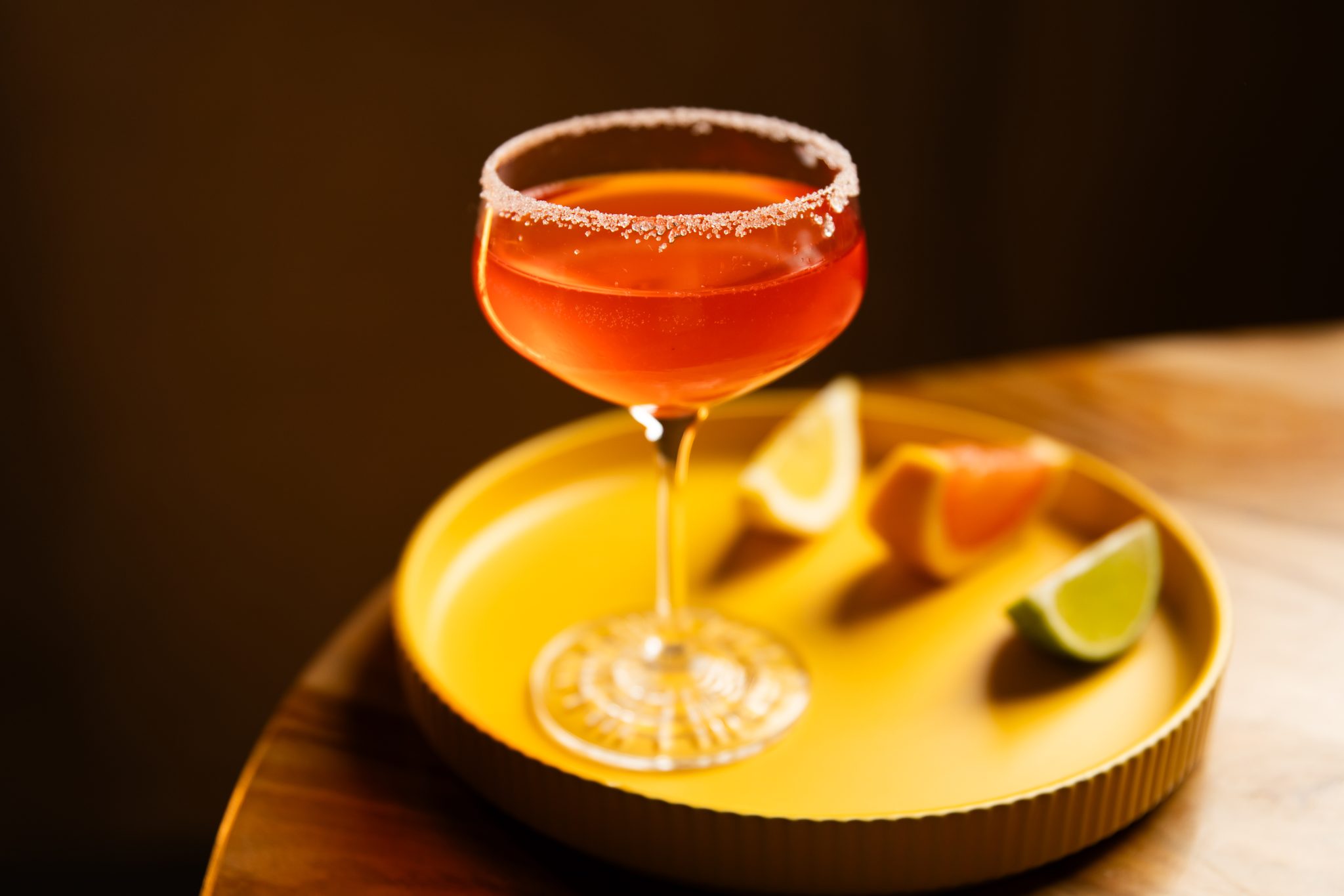 A side shot of a Bald Eagle drink in a coupe glass on a yellow tray with a lemon wedge, a lime wedge, and a pink grapefruit wedge on the side