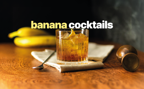 A shot of a banana cocktail in an Old Fashioned glass on a wooden surface surrounded by two bananas, a bar spoon, a jigger and two white cloth