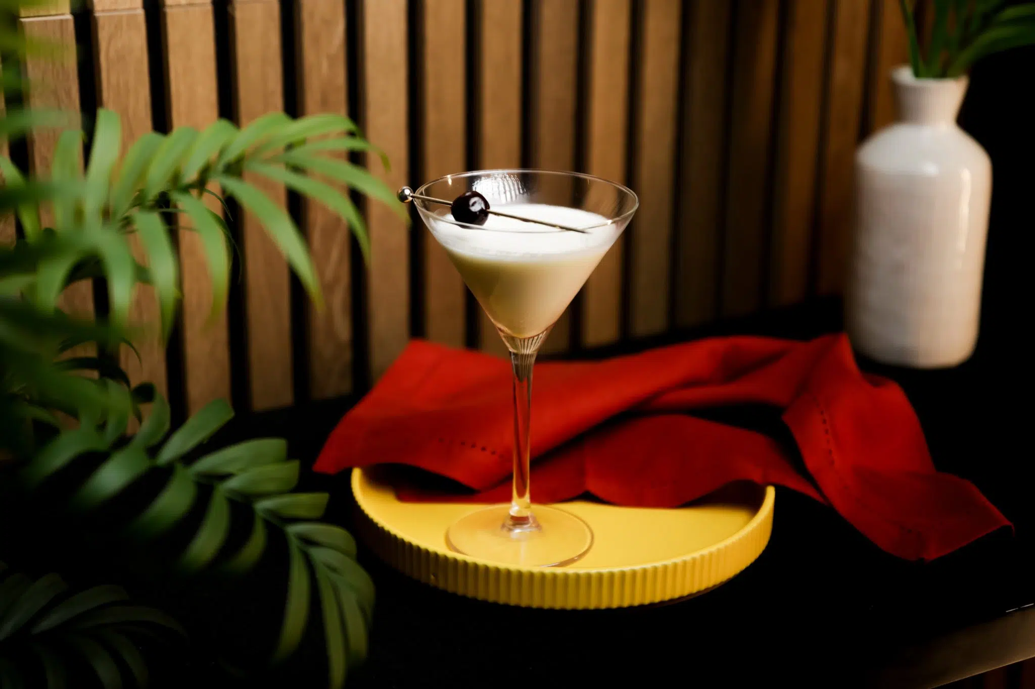 A side shot of a Banana Split Martini cocktail in a martini glass on a yellow tray with a red cloth on the side and a white bottle