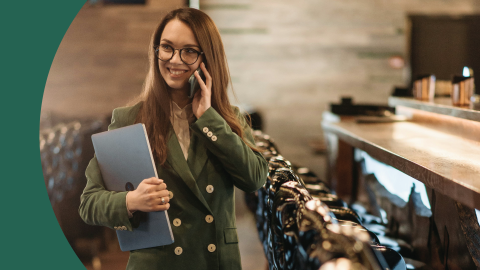 A shot of a woman wearing a green suit talking with the phone and holding a laptop with the other hand in front of a bar table