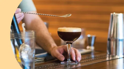 A shot of a bartender holding a bar spoon over a coupe glass with a cocktail on a bar table