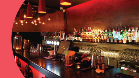 A shot of a black bar table with two shelves behind filled with spirits bottles, and different types of glasses