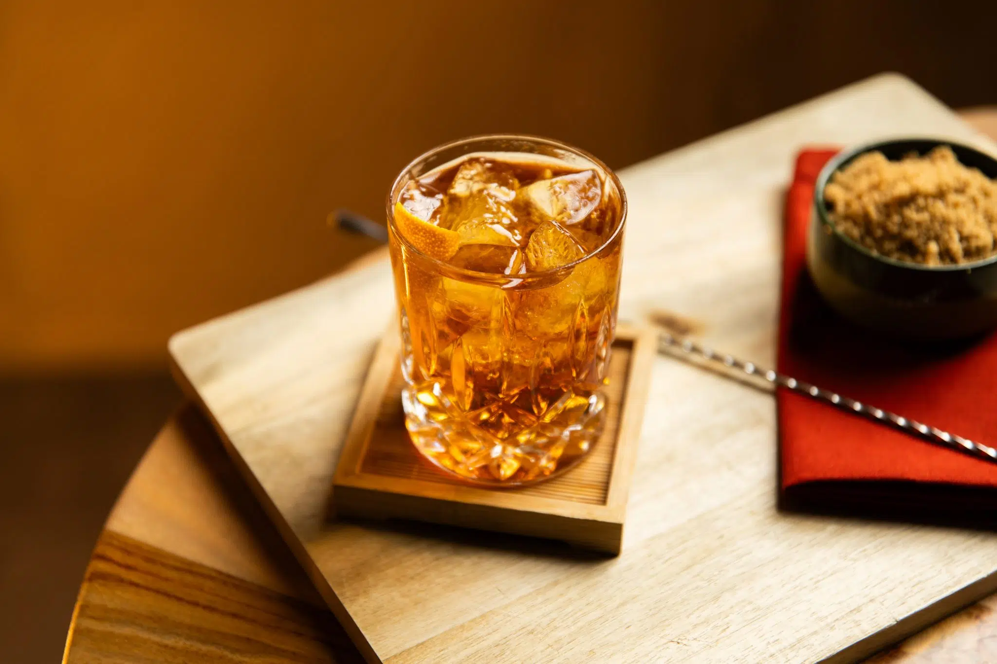 A side shot of a Bitter in Brazil cocktail in an old fashioned glass on a wooden coaster placed on a wooden board surrounded by a bar spoon and a bowl with Demerara sugar