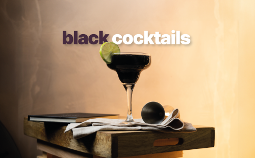 A black cocktail in a margarita glass on a wooden tray surrounded by a jigger and a white cloth