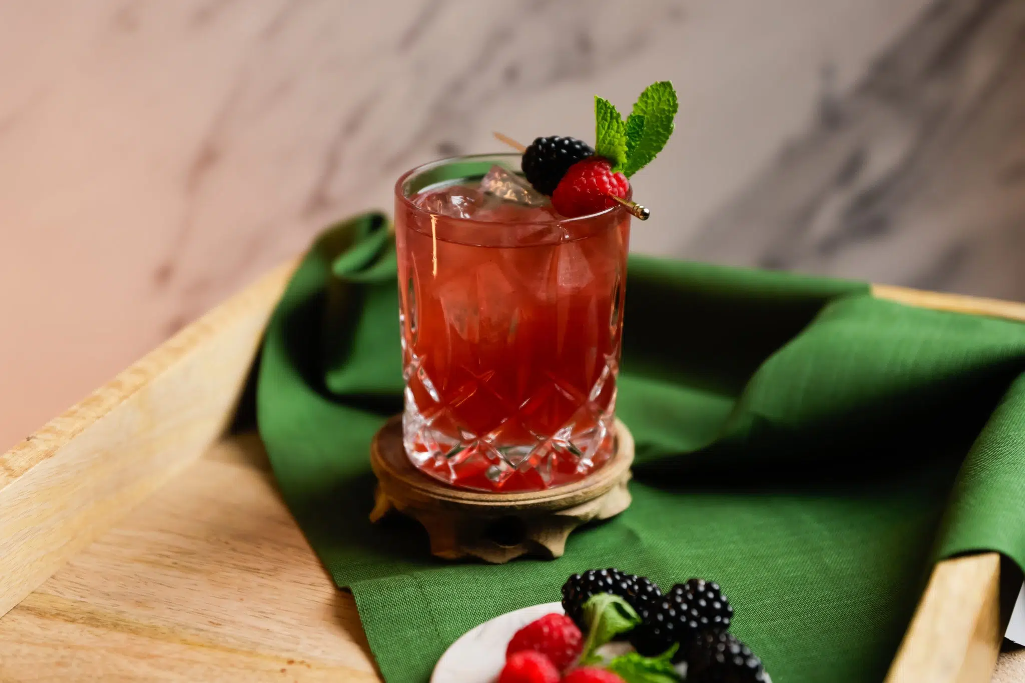 A side shot of a Black Eye drink in an old fashioned glass on a wooden coaster on a wooden surface surrounded by a green cloth and a white coaster with raspberries and blackberries