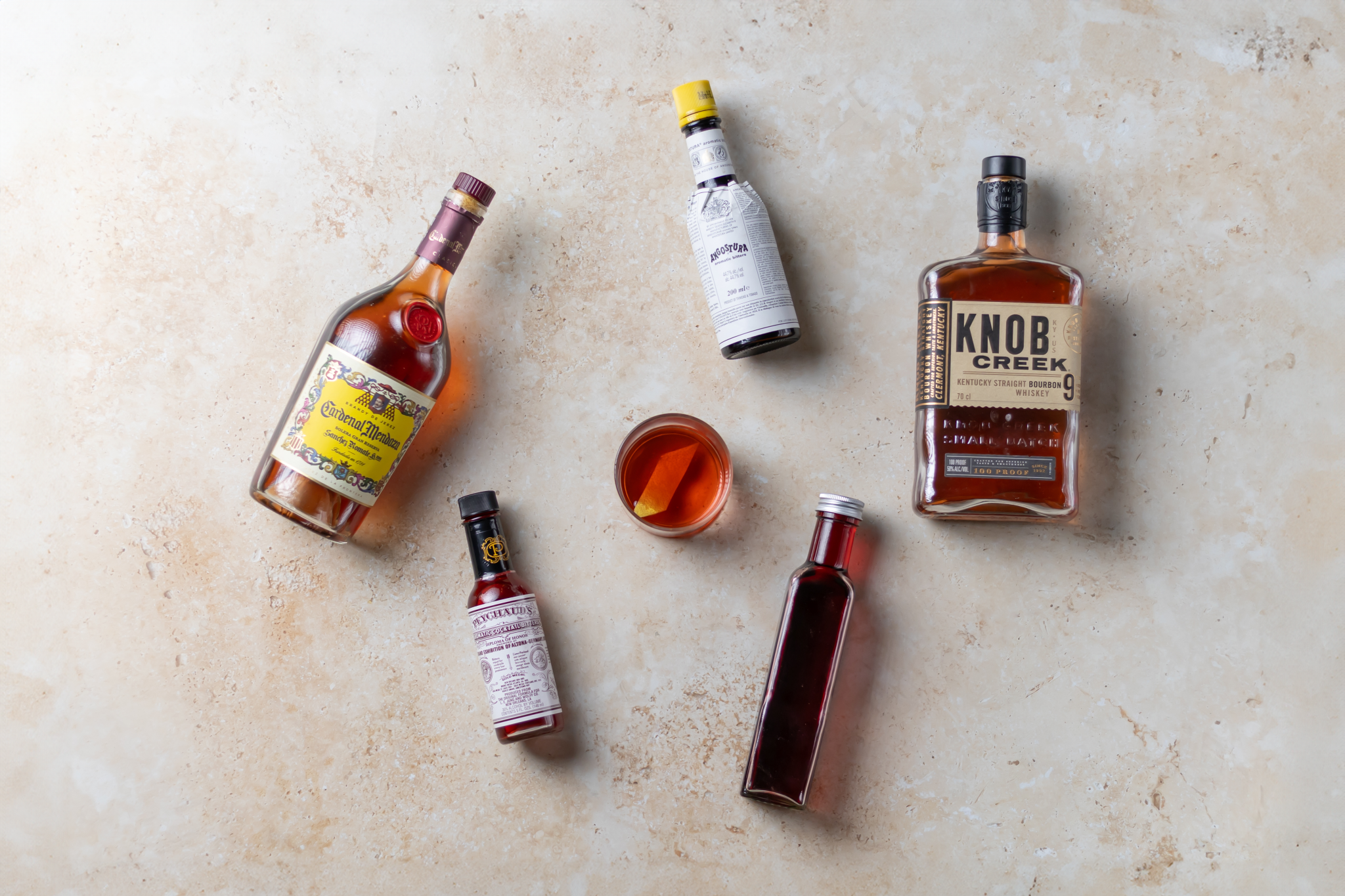 A Black Rose cocktail, shot from above, in an old fashioned glass on a beige surface surrounded by Angostura bitters, Peychaud's bitters, grenadine, bourbon, and cognac