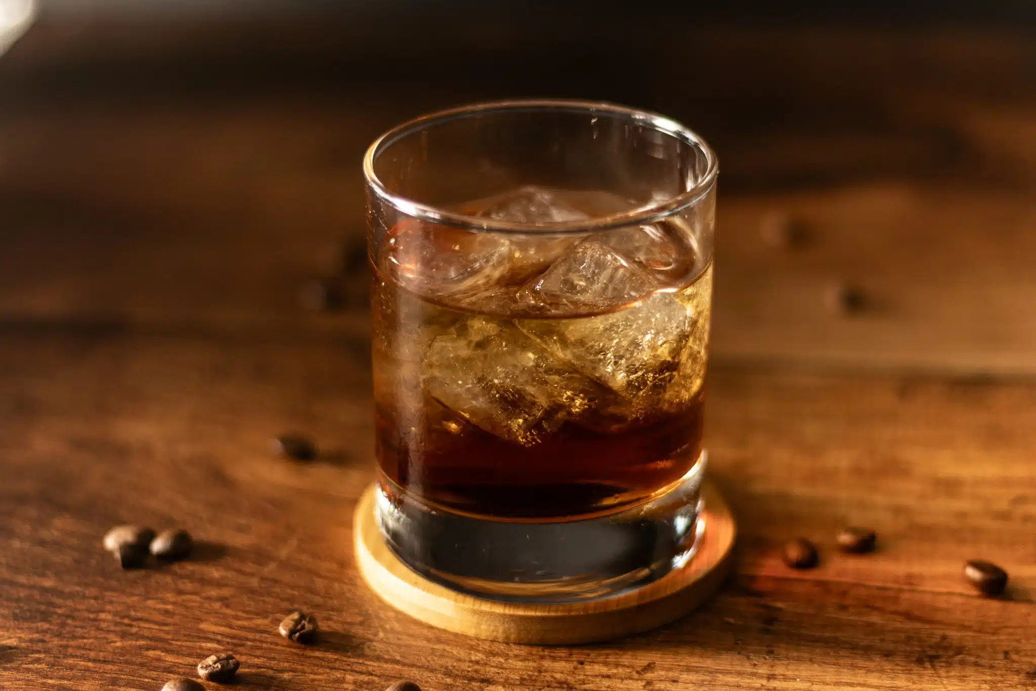 A side shot of a Black Russian cocktail in an old fashioned glass on a light wooden coaster placed on a drak wooden table surrounded by coffee beans