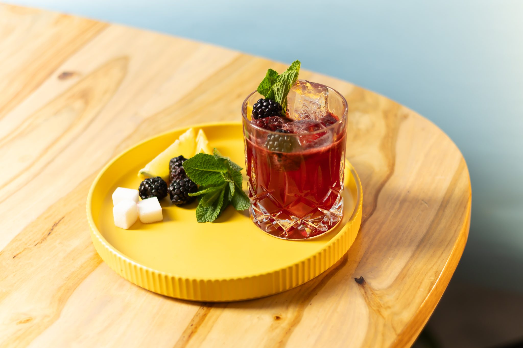 A side shot of a Blackberry Bourbon Smash cocktail in an old fashioned glass on a yellow tray placed on a wooden table surrounded by three sugar cubes, three blackberries, two lemon wedges, and a mint sprig