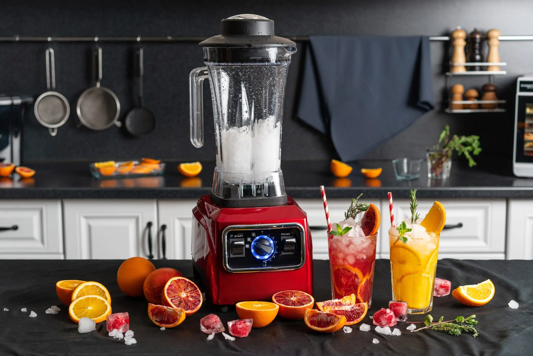 A blender with a transparent glass filled with crushed ice and a red base, placed on a black surface in a kitchen with two drinks on the side and fruits and ice around