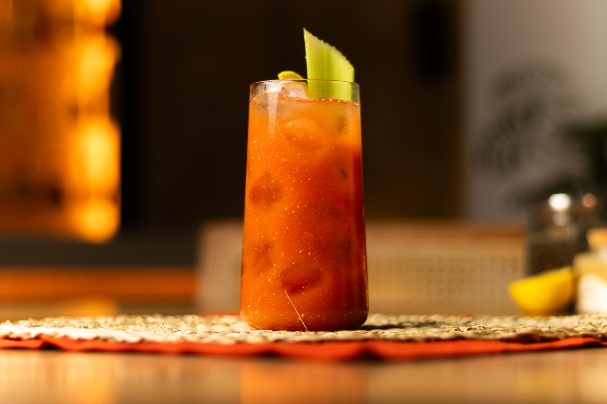 A side shot of a Bloody Maria in a highball glass on a white and red placemat placed on a wooden table with a blurry background.