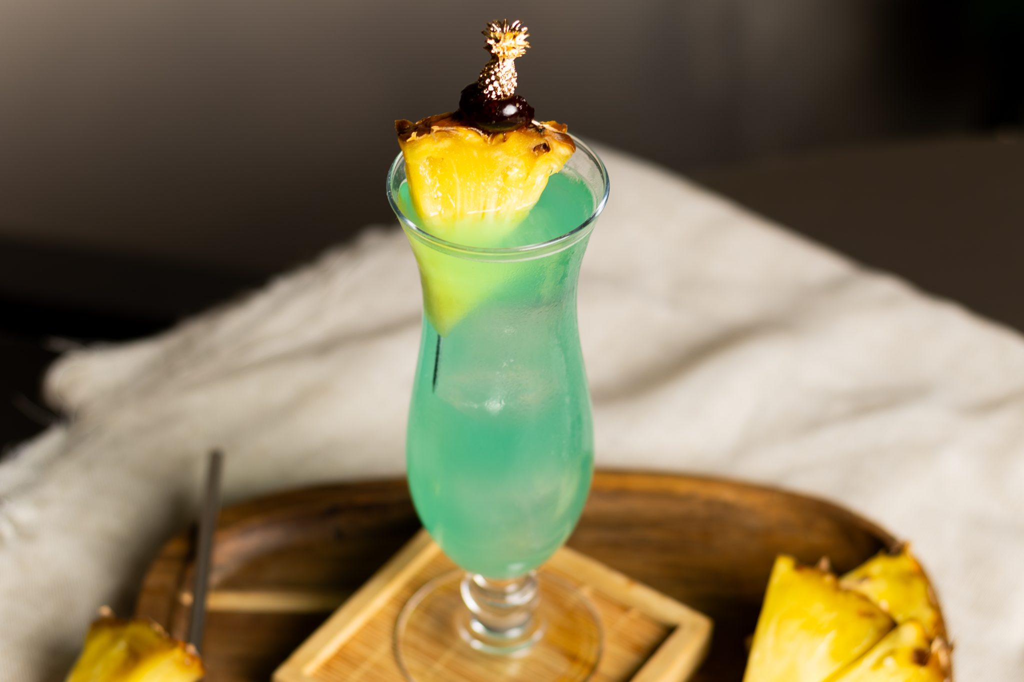 A side shot of Blue Hawaii cocktail in a hurricane glass on a wooden coaster placed on a wooden tray surrounded by white cloth two pineapple slices.
