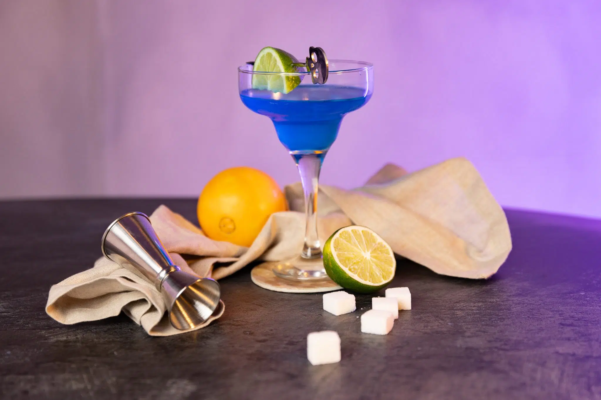 A side shot of a Blue Margarita cocktail in a margarita glass on a dark grey surface suttounded by an orange, a lime, a jigger, five sugar cubes and a beige cloth