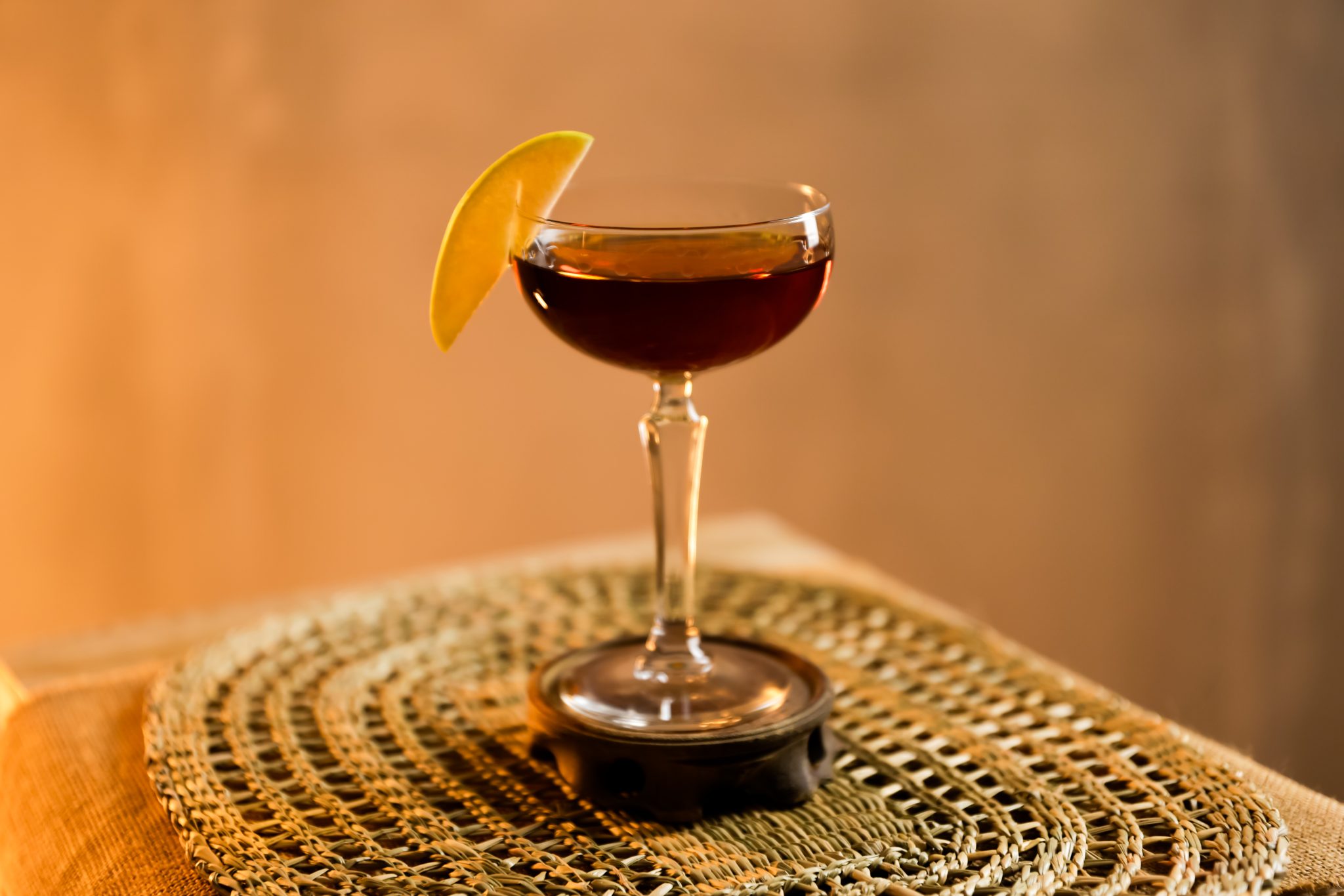 A side shot of a Bolero cocktail in a coupe glass on a wooden coaster placed on a straw placemat in front of a brown background