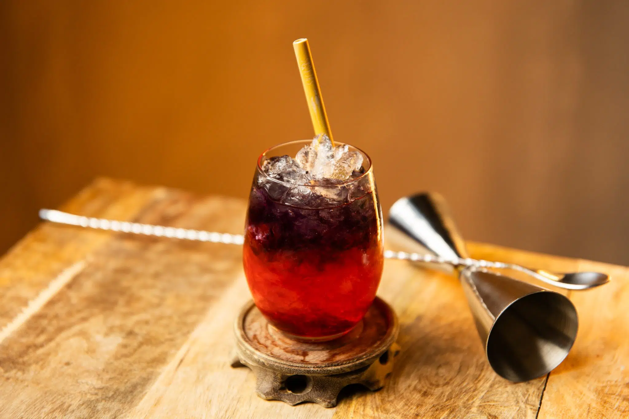 A side shot of a Bomb Pop drink in a highball glass on a wooden coaster and board with a jigger and a bar spoon behind.