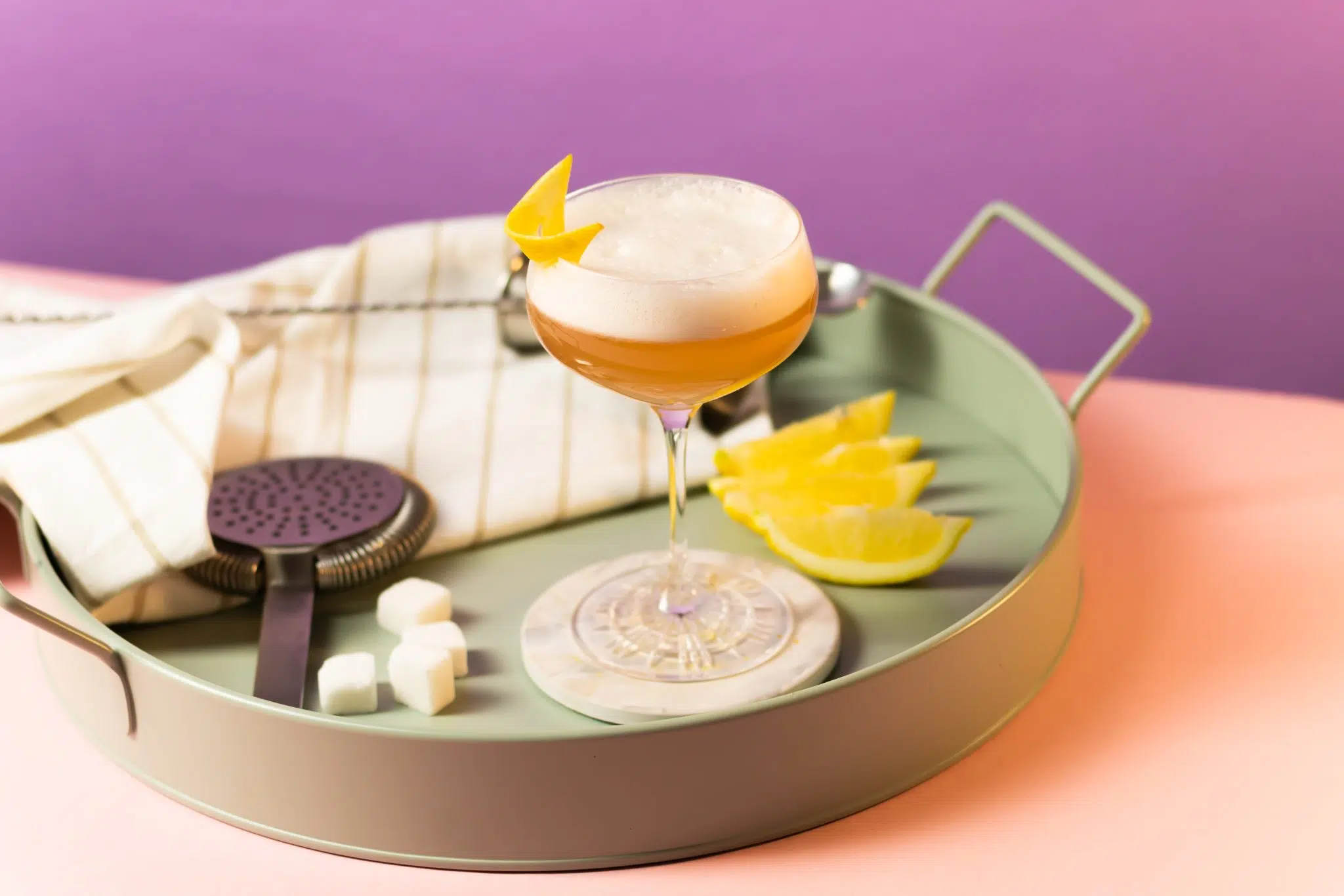 A side shot of a Boston Sour cocktail in a cocktail glass on a white coaster placed on a turquoise tray surrounded by a a cocktail strainer, four lemon wedges, four sugar cubes, a bar spoon, and a white cloth