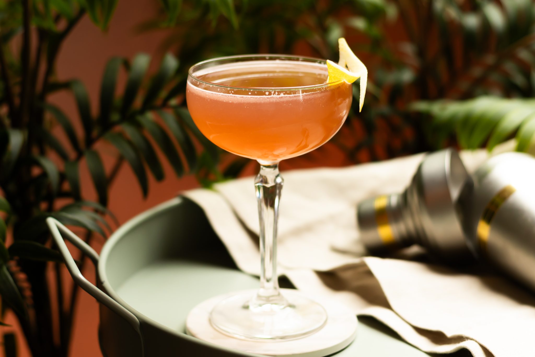 A side shot of a Bourbon Daisy cocktail in a coupe glass on a white coaster placed on a turquoise tray with a shaker and a white cloth on the side