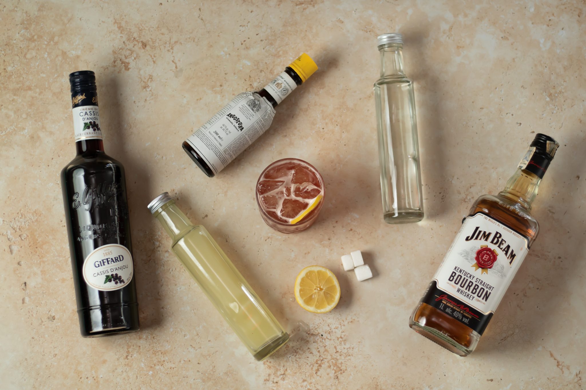 A Bourbon Renewal cocktail, shot from above, in an old fashioned glass on a beige surface surrounded by Bourbon, lemon juice, Creme de Cassis, Rich Syrup, Angostura aromatic bitters, sugar cubes and lemon