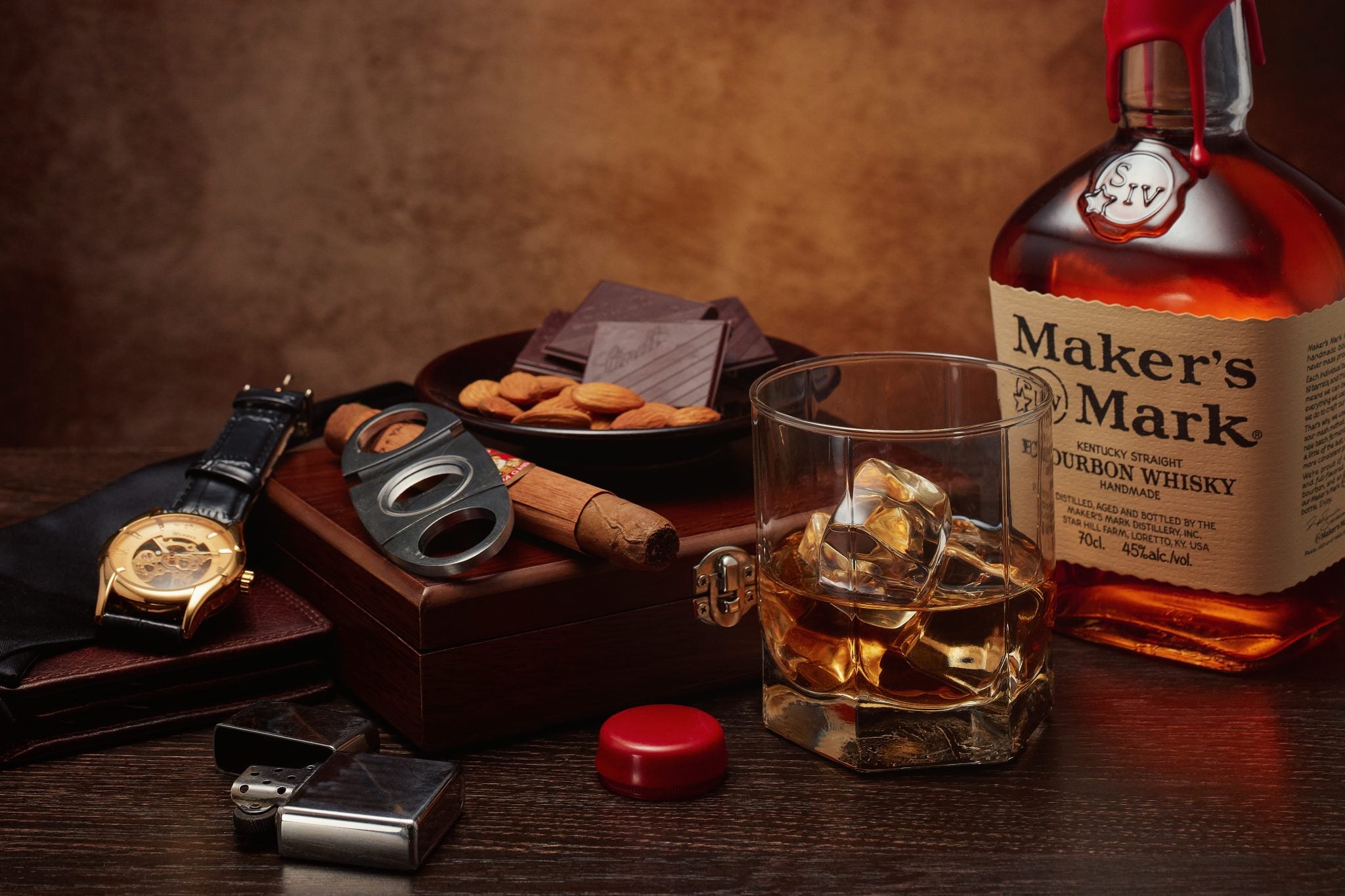 A shot of a bottle and an Old Fashioned glass of Makers Mark Kentucky Bourbon Whiskey on a wooden surface surrounded by a watch, a lighter, a cutter, a cigar, a brown wallet, a plate with almonds and dark chocolate and a wooden box