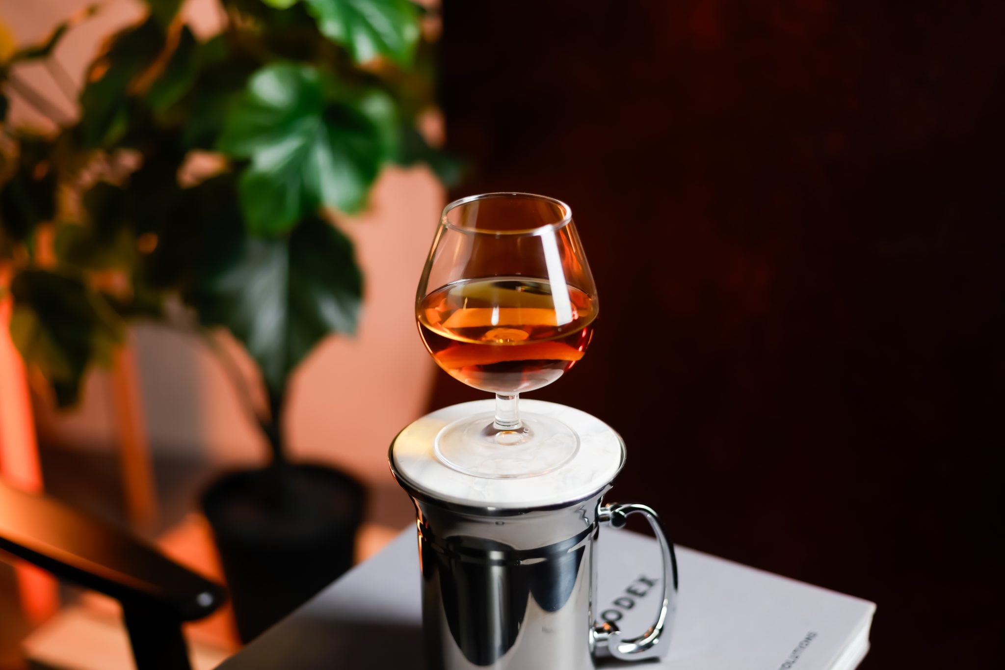 A side shot of a Brandy Blazer cocktail in a cognac glass on a white coaster on a metal blazer mug, in front of a plant