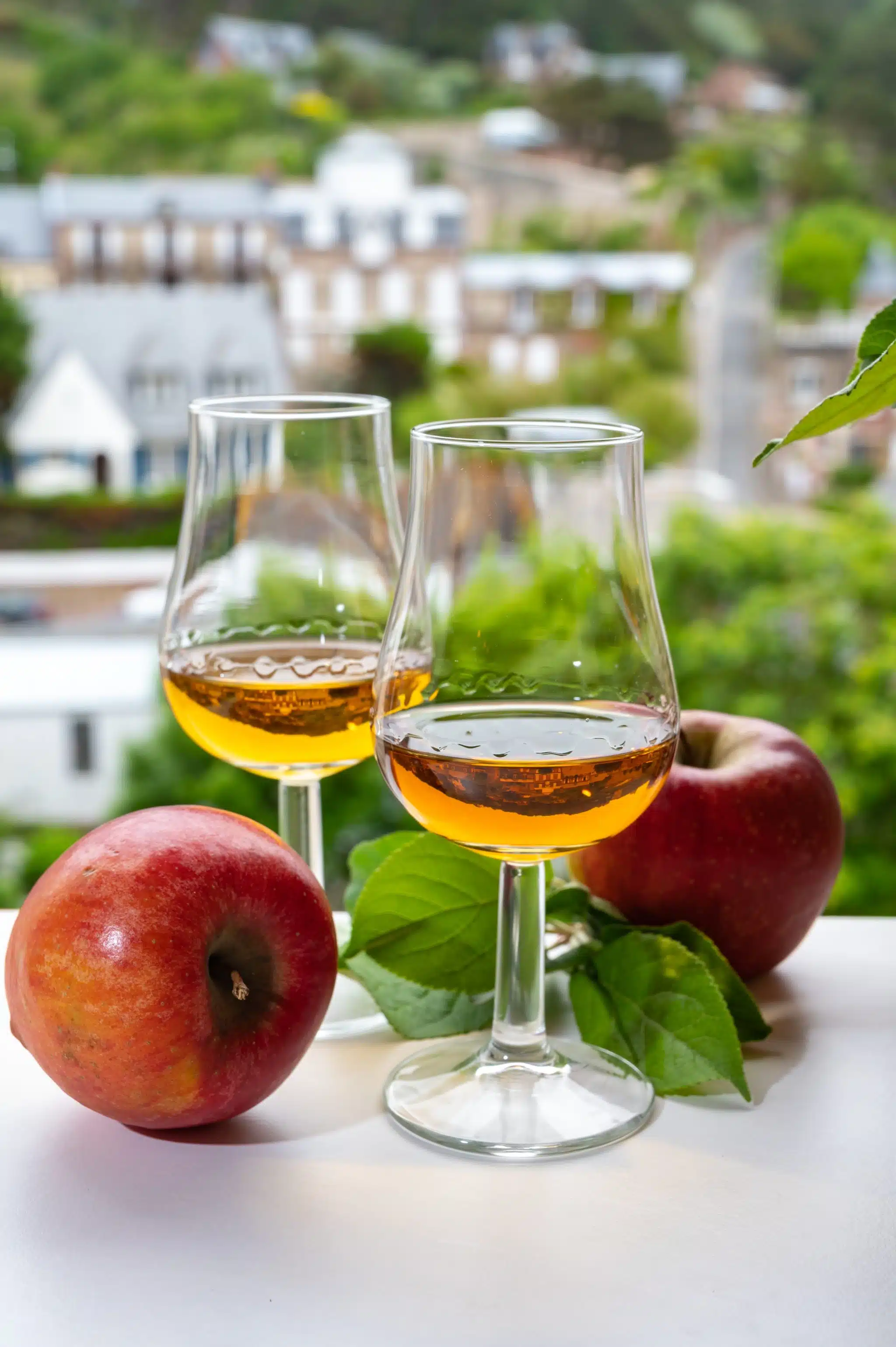 Two glasses of Calvados brandy on a balcony with red apples, with a blurred village scene on the background.