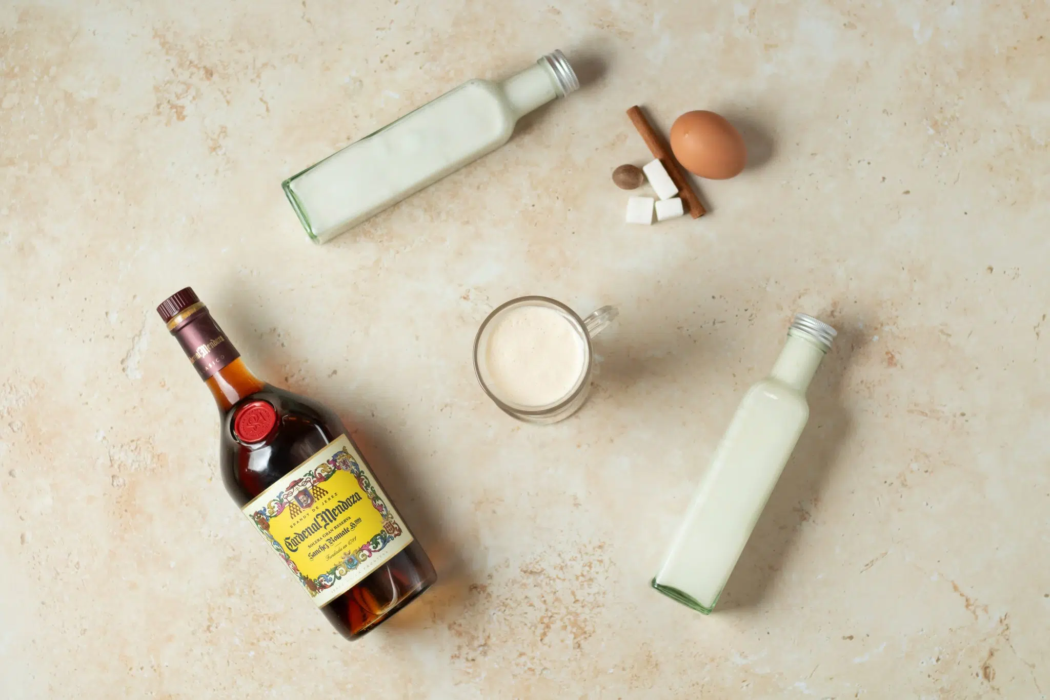 A Brandy Eggnog cocktail, shot from above, in a mug on a beige surface surrounded by Brandy, whole milk, heavy cream, sugar cubes, cinnamon sticks, nutmeg and egg.